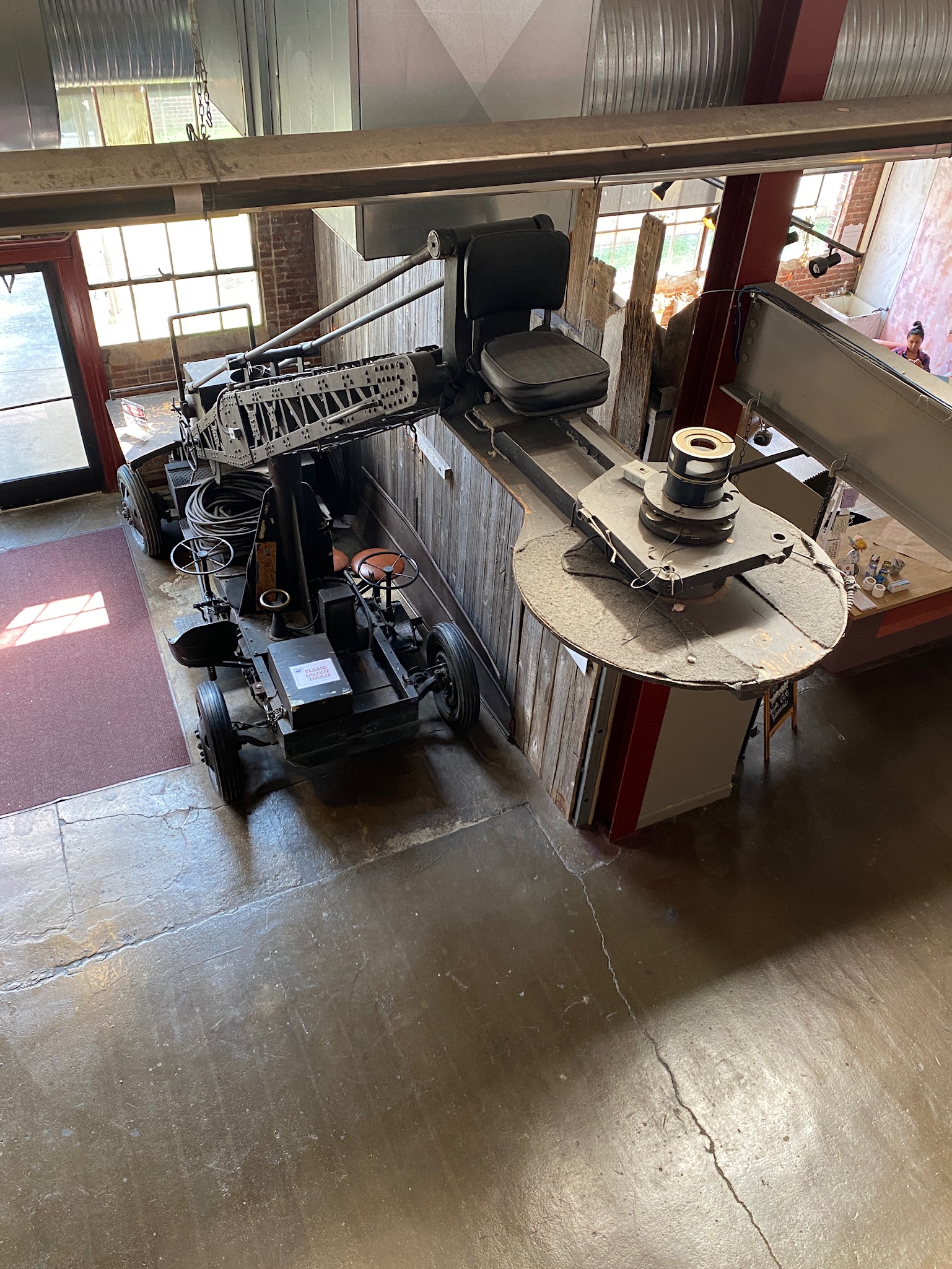A large vintage film camera crane is positioned indoors near a window, with its arm extended over a railing on a polished concrete floor.