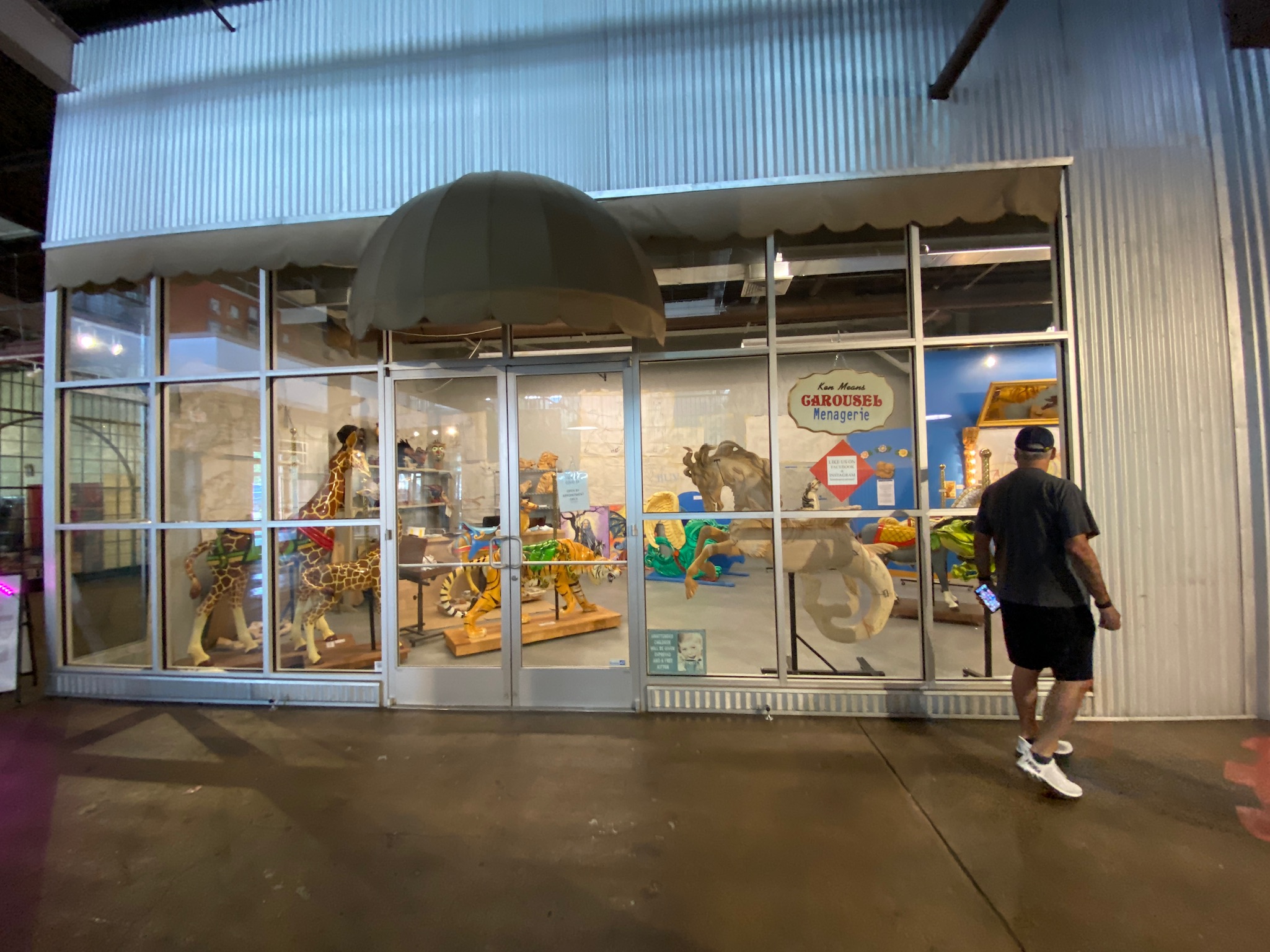 A person walks past a storefront with large glass windows displaying wooden animal sculptures and toys under an awning inside a building.