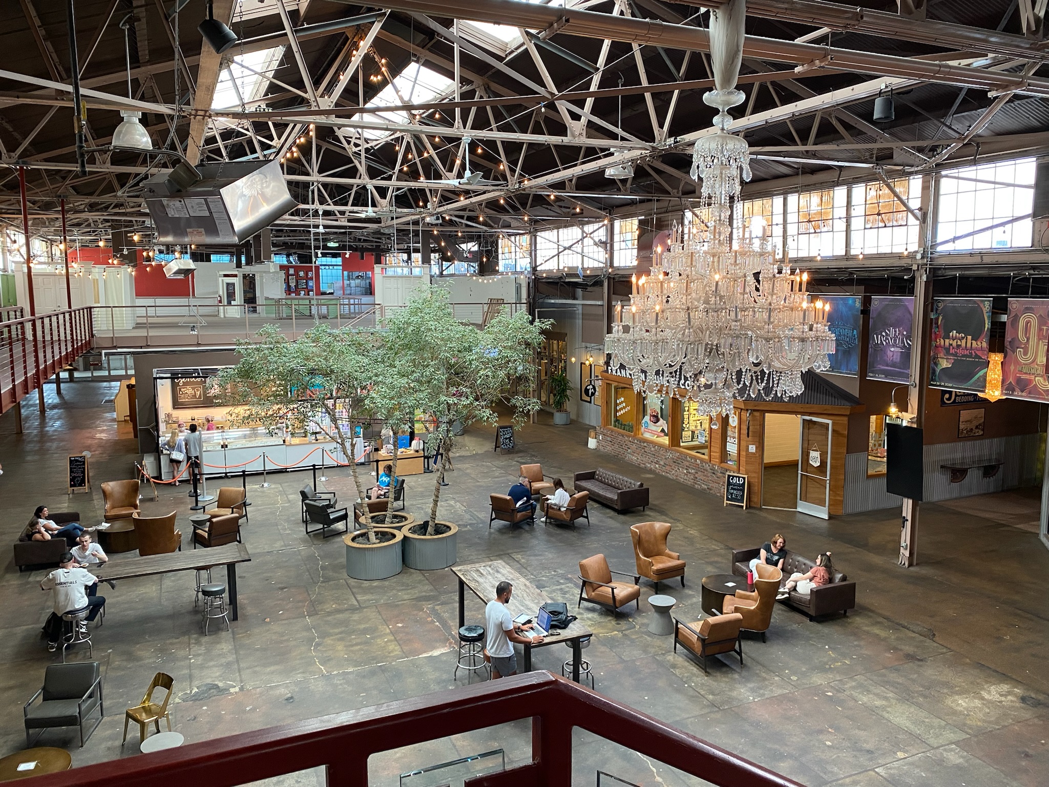 Spacious indoor area with scattered lounge seating, central trees in planters, a large chandelier, and people seated or walking around under a high industrial ceiling.