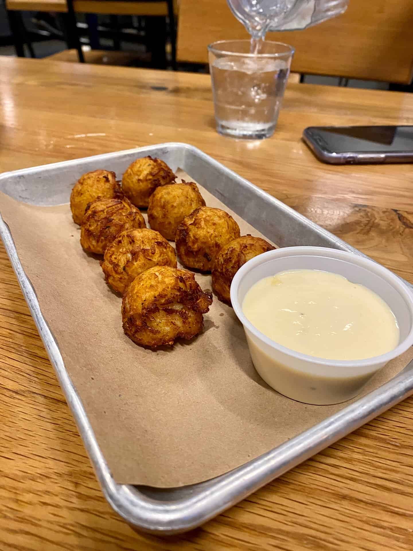 A tray with eight fried round snacks next to a small container of dipping sauce. A glass is being filled with water in the background.