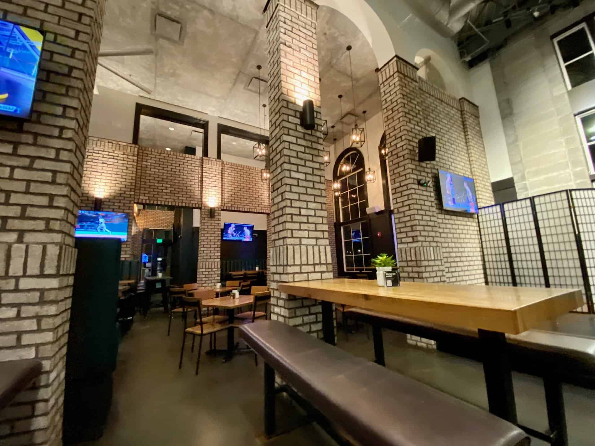 Spacious restaurant interior with brick walls, wooden tables, and TV screens. Large windows and industrial ceiling create an open atmosphere. Small plant on a table adds a touch of greenery.