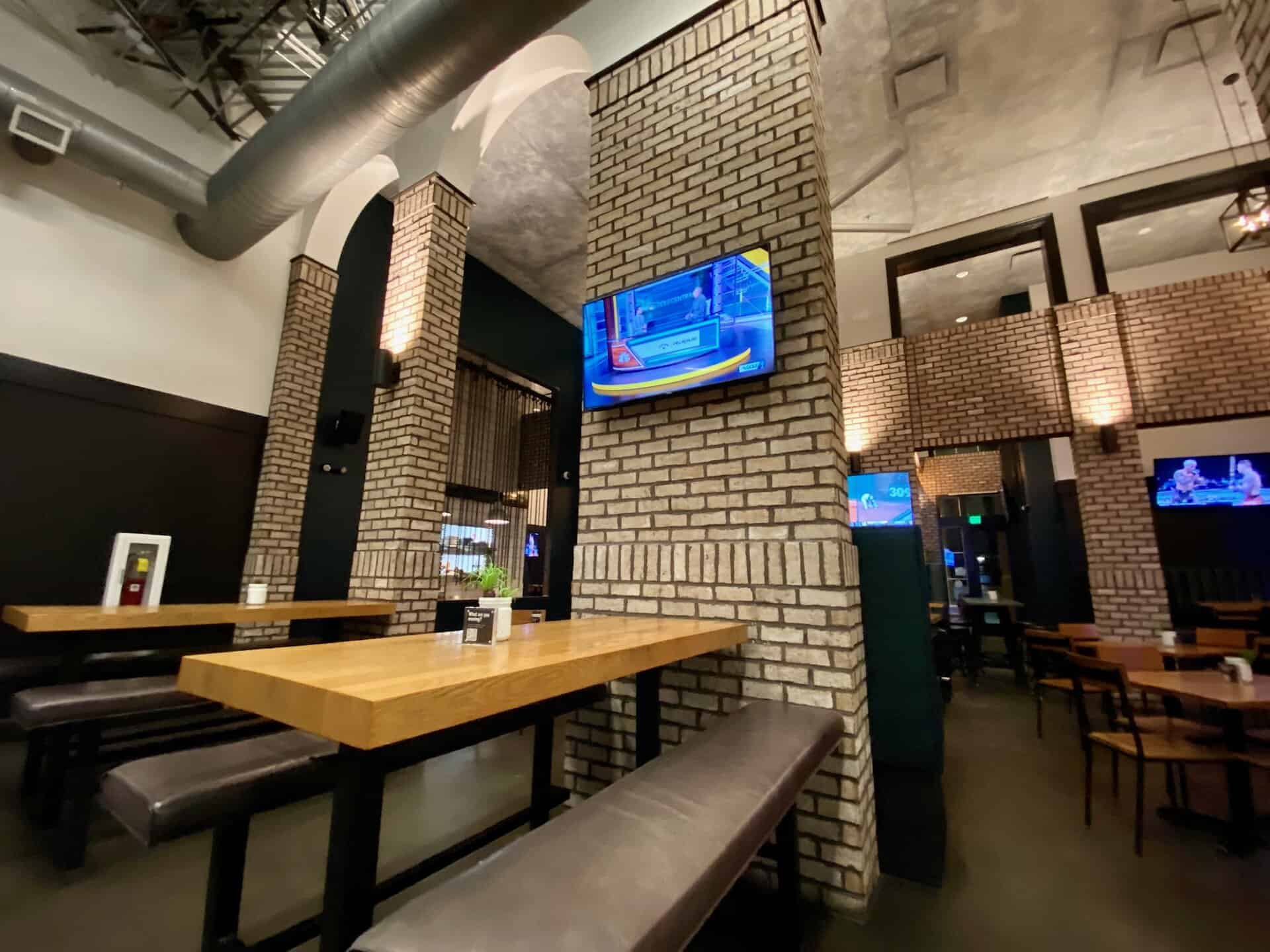 Interior of a modern sports bar with brick walls, wooden tables, and benches. Multiple TVs are mounted showing various sports channels. The ceiling features exposed ductwork.