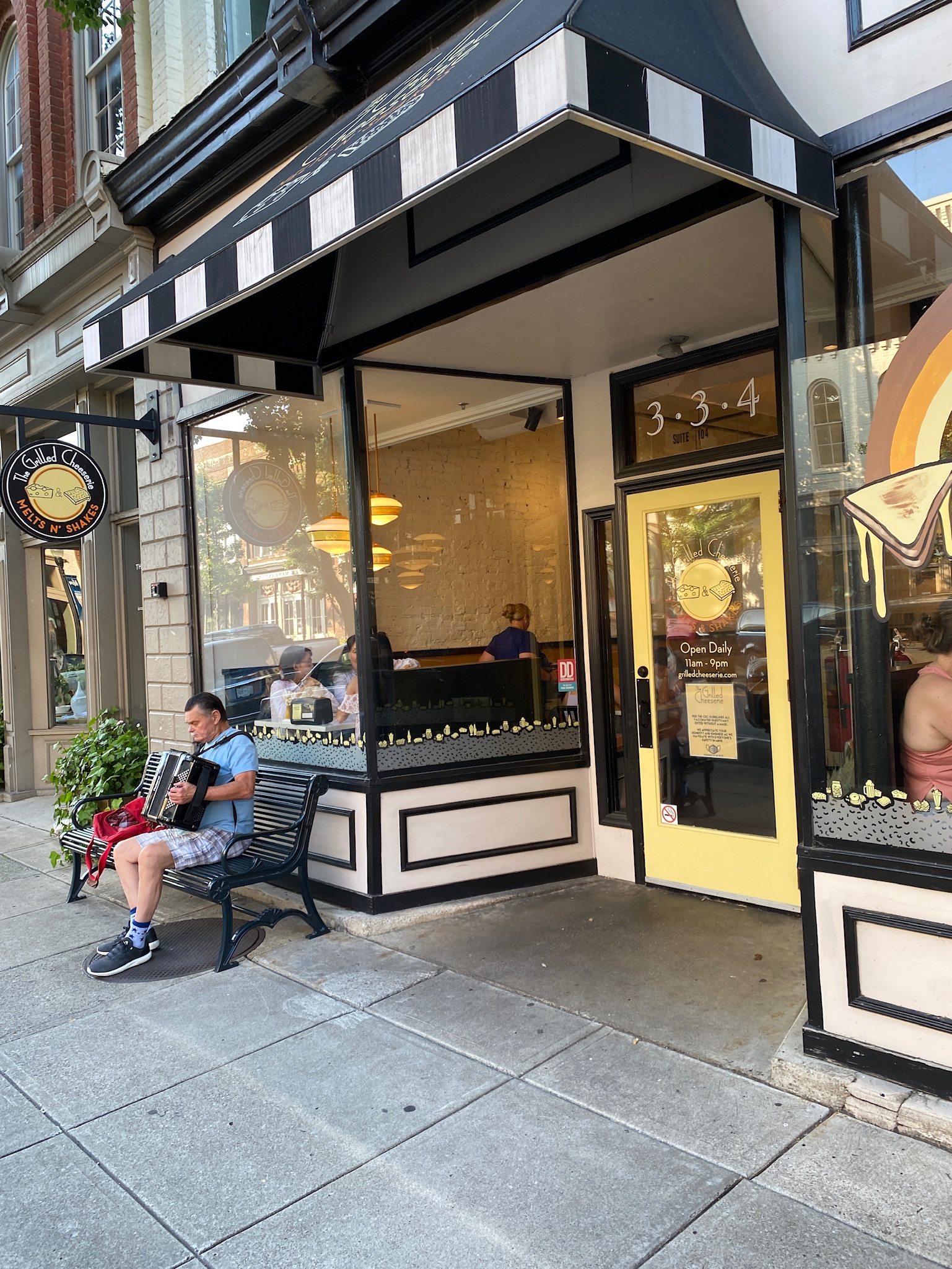 A man sits on a bench playing an accordion outside a yellow and black storefront with large windows and people inside.