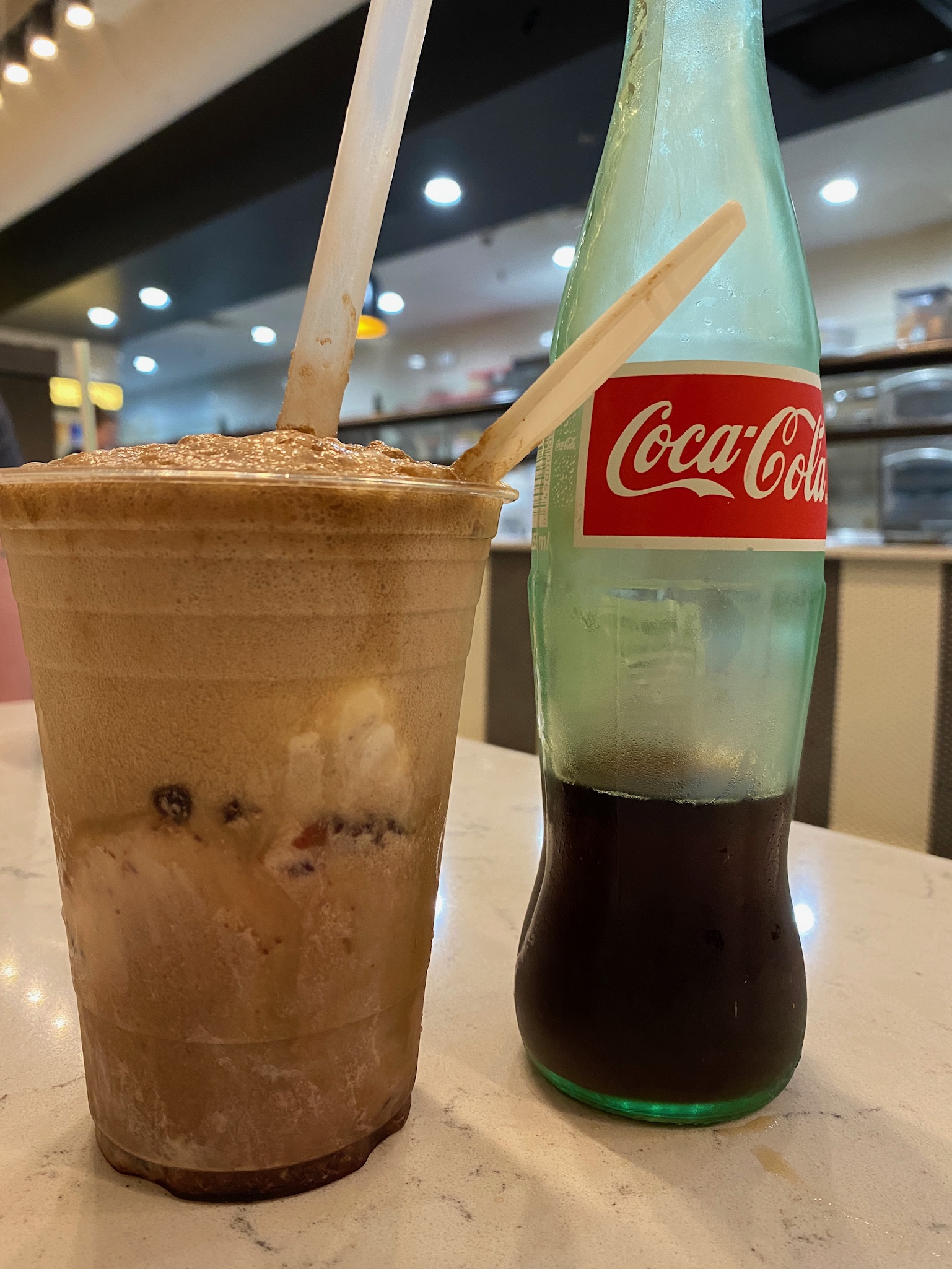 A plastic cup with a Coca-Cola float and two straws sits next to a partially filled glass bottle of Coca-Cola on a marble table in a restaurant.