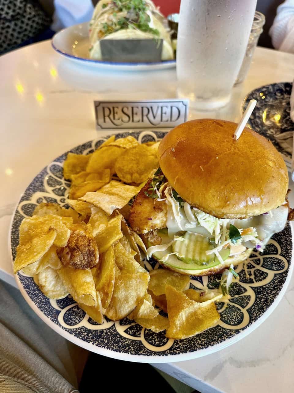 A Nashville-style chicken sandwich with coleslaw is elegantly presented on a decorative plate, served alongside crispy potato chips. A "Reserved" sign sits next to a refreshing glass of water on the table, completing this Southern dining experience.