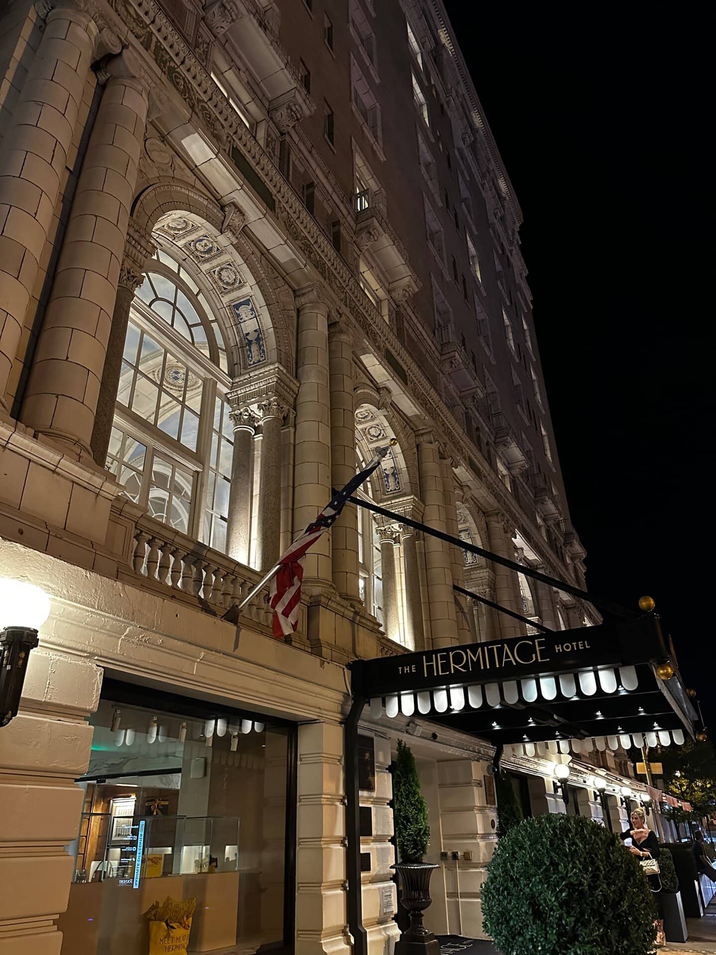 Night view of The Hermitage Hotel entrance in Nashville, featuring ornate architecture with illuminated windows and an American flag.