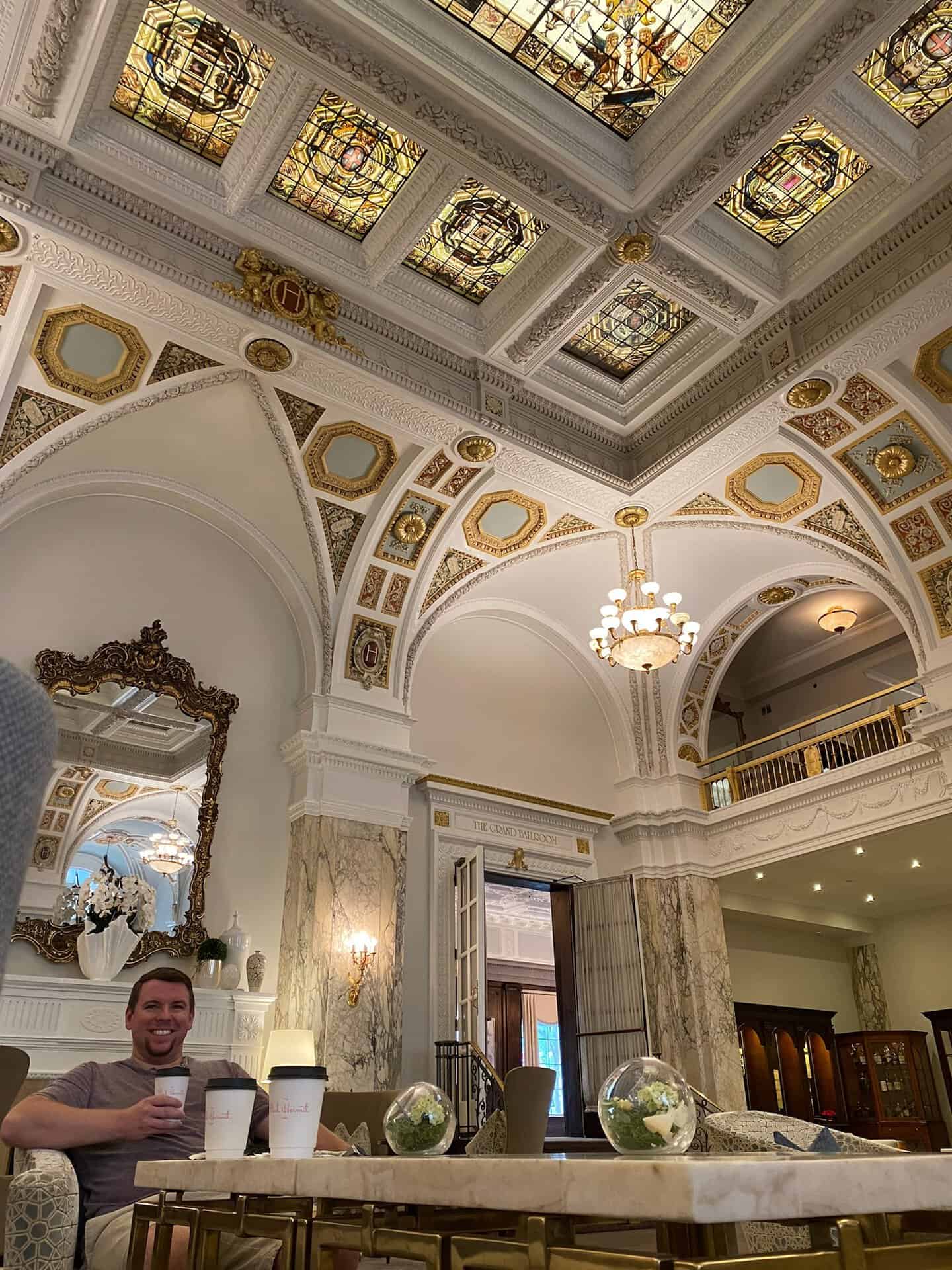 A man sits with coffee in an ornate Nashville lobby featuring high ceilings, stained glass, and decorative arches, next to an elegant mirror and marble columns.