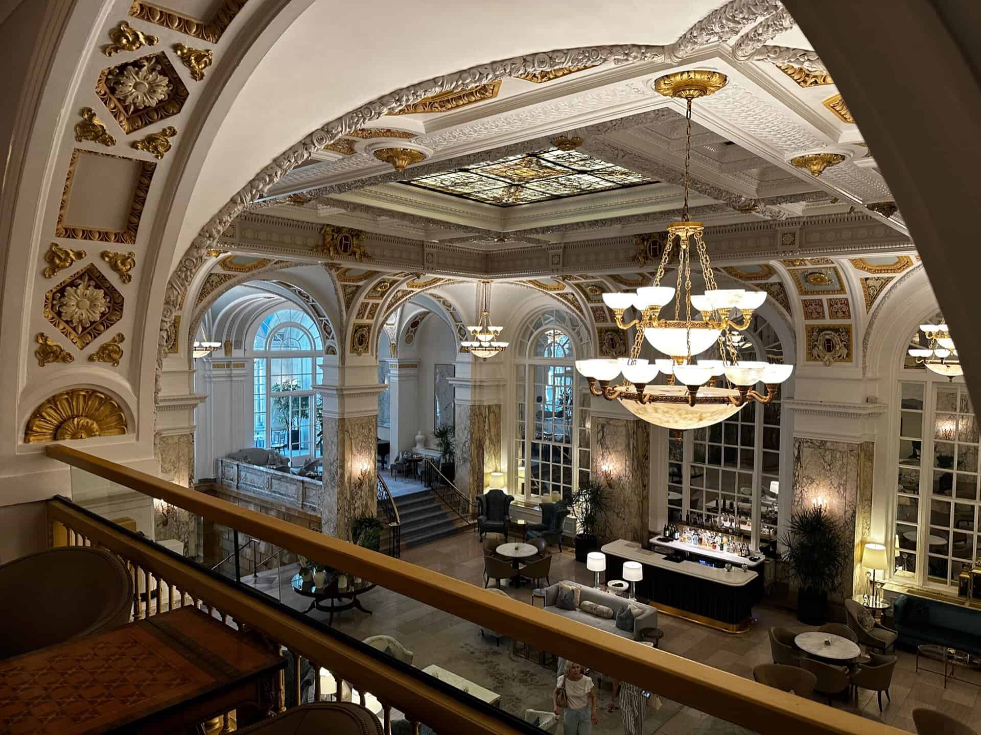 Elegant hotel lobby with ornate arches, chandeliers, and a stained glass ceiling, echoing Nashville's vibrant charm. The spacious room features large windows and decorative molding, creating a luxurious atmosphere complemented by a welcoming bar.