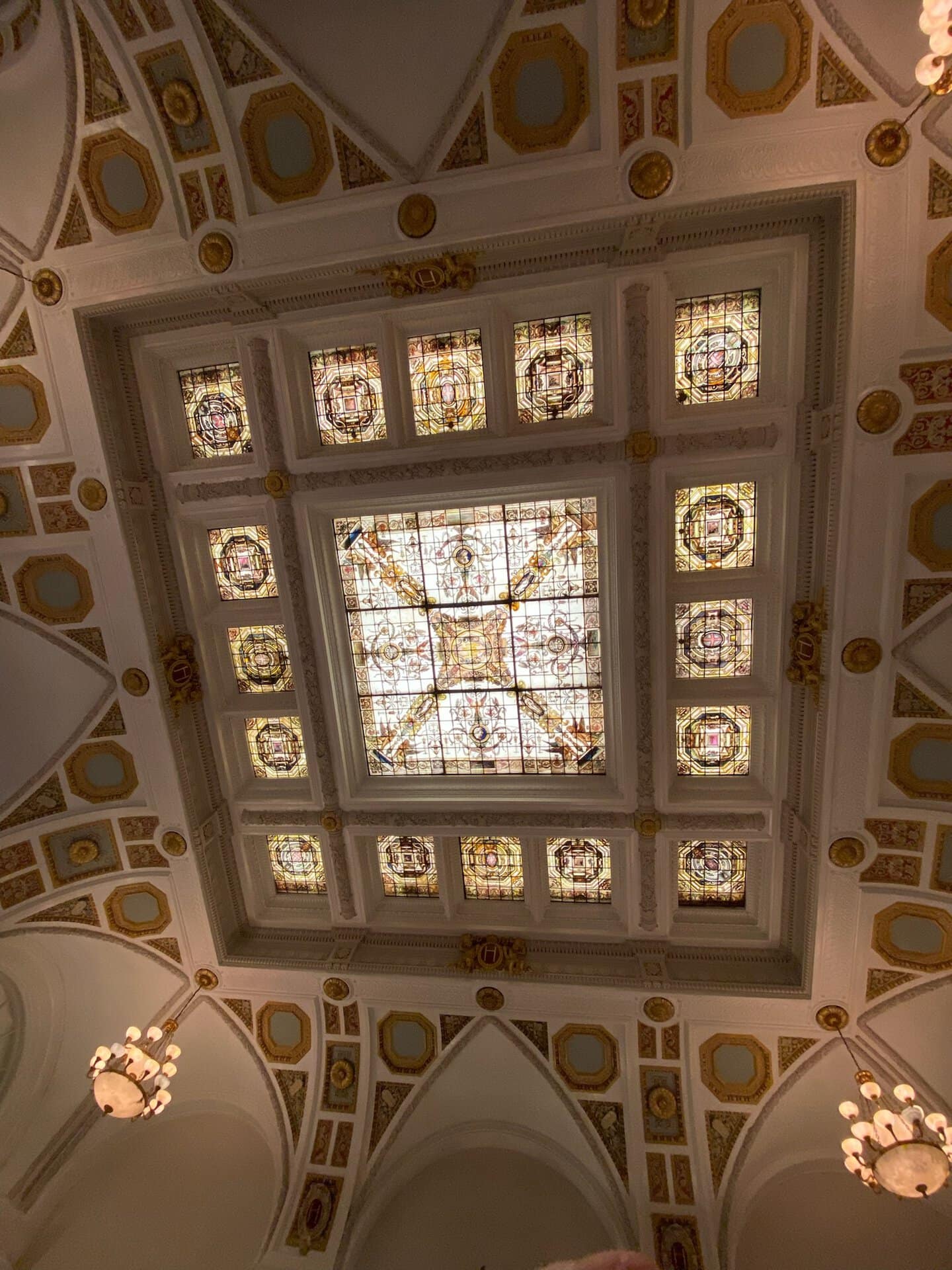 The ornate ceiling in Nashville dazzles with stained glass and decorative molding, showcasing a central square panel surrounded by chandeliers and intricate architectural details.