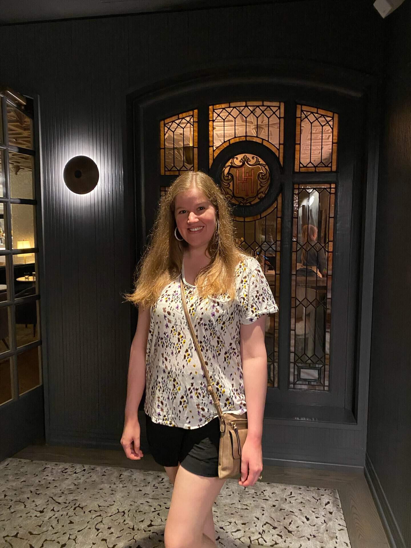 A person with long hair stands indoors in front of a decorative stained glass window, wearing a white patterned shirt and black shorts, smiling at the camera—a little hint of Nashville charm in their style.