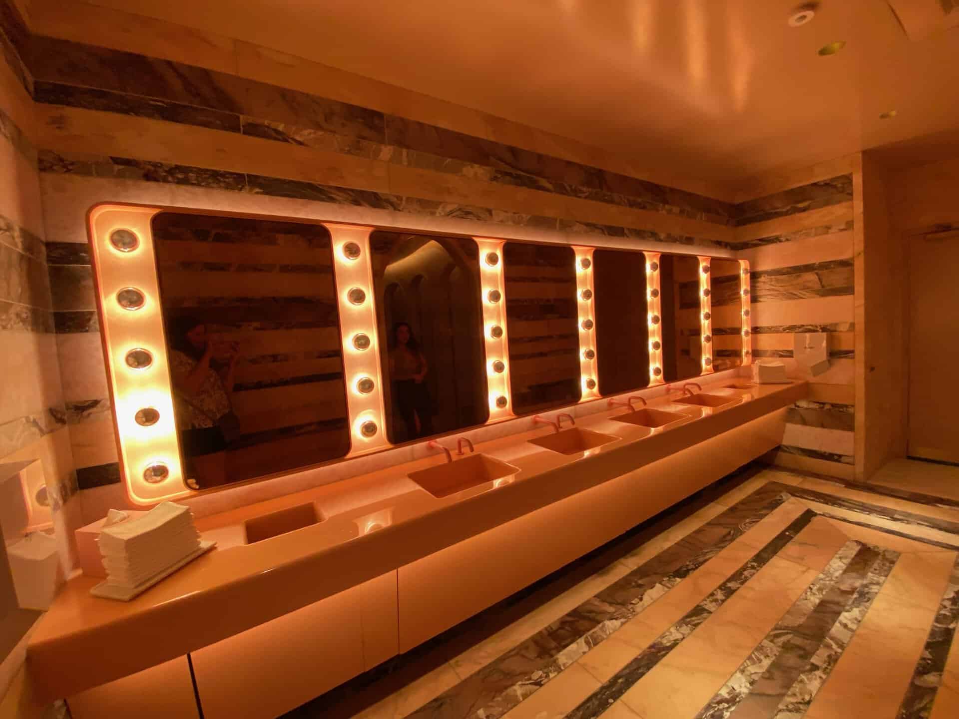 A luxurious Nashville bathroom with marble walls, illuminated by a large mirror surrounded by round lights, features multiple sinks and a stack of paper towels on the counter.