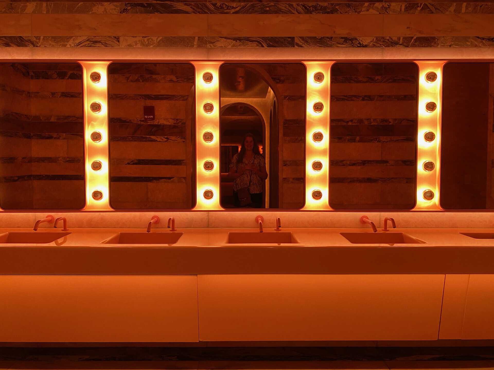 A dimly lit restroom in Nashville boasts a row of illuminated mirrors above sinks, featuring glowing circular lights and a person faintly visible in the mirror reflection.