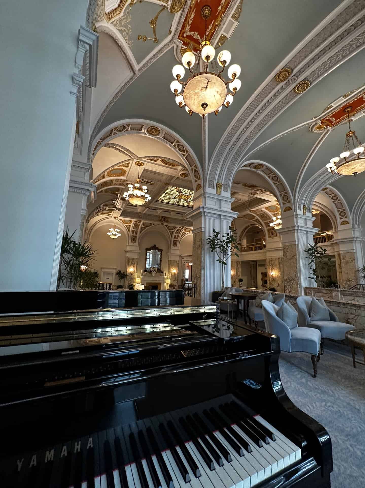 Grand ornate lobby with chandeliers and arched ceilings, featuring a black Yamaha piano in the foreground. Elegant seating and detailed architectural elements enhance the luxurious ambiance, reminiscent of Nashville's opulent charm.