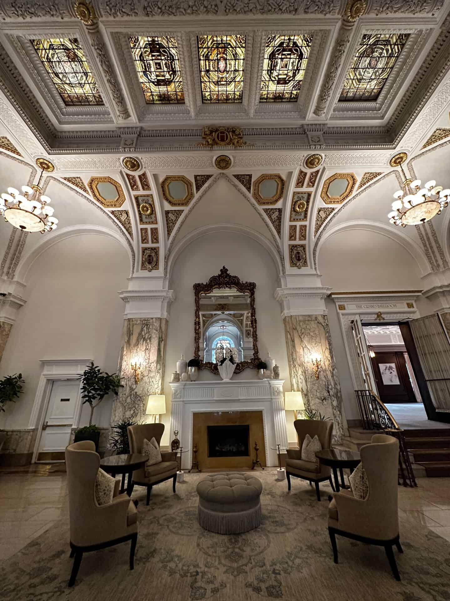 The elegant Nashville hotel lobby boasts an ornate ceiling, stained glass, and chandeliers. A central fireplace is flanked by armchairs and a large mirror, while plush carpeting and potted plants add to the sophisticated decor.