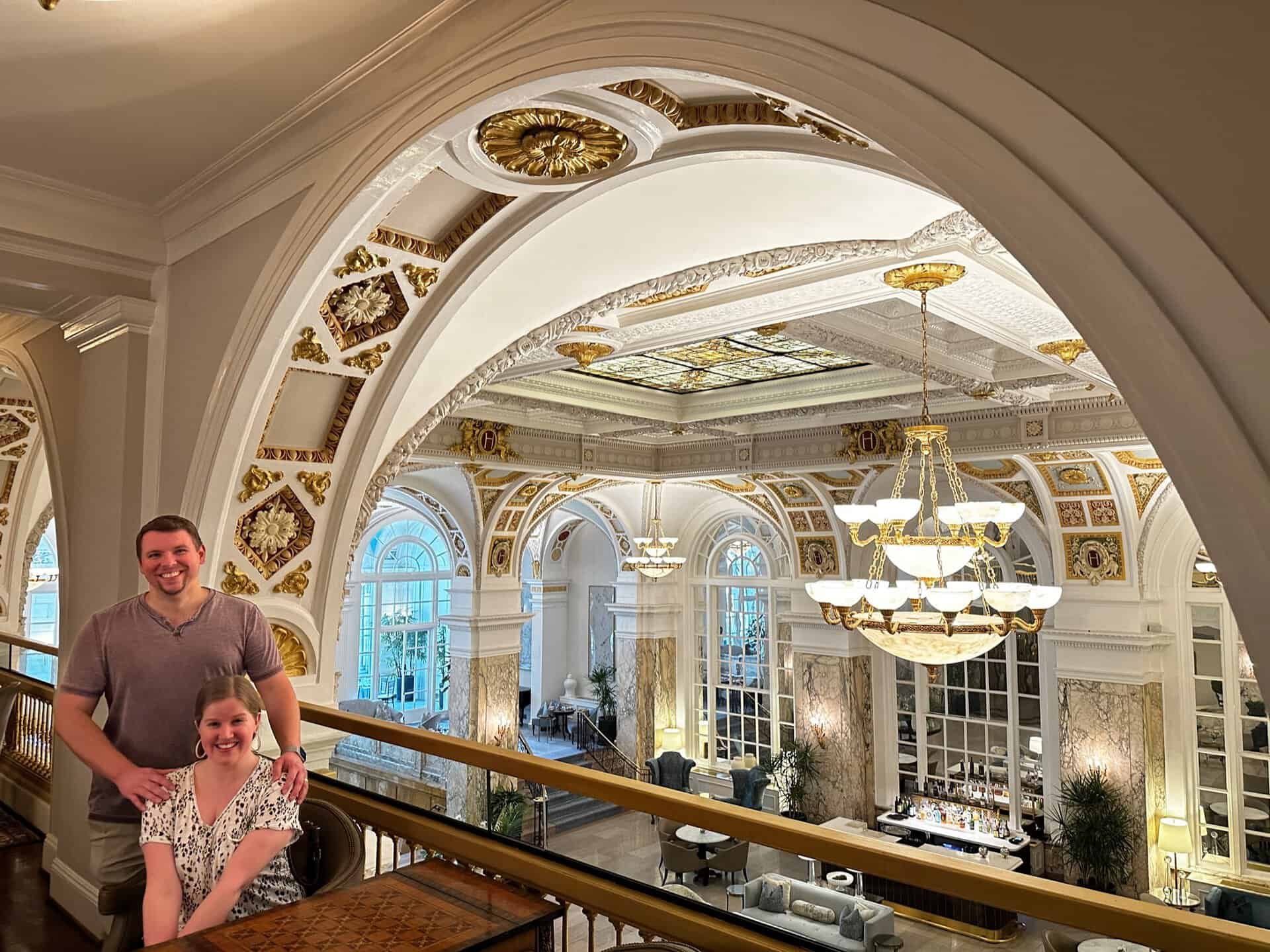 A smiling couple poses in a grand interior reminiscent of Nashville's opulent charm, featuring ornate arches, chandeliers, and large windows.