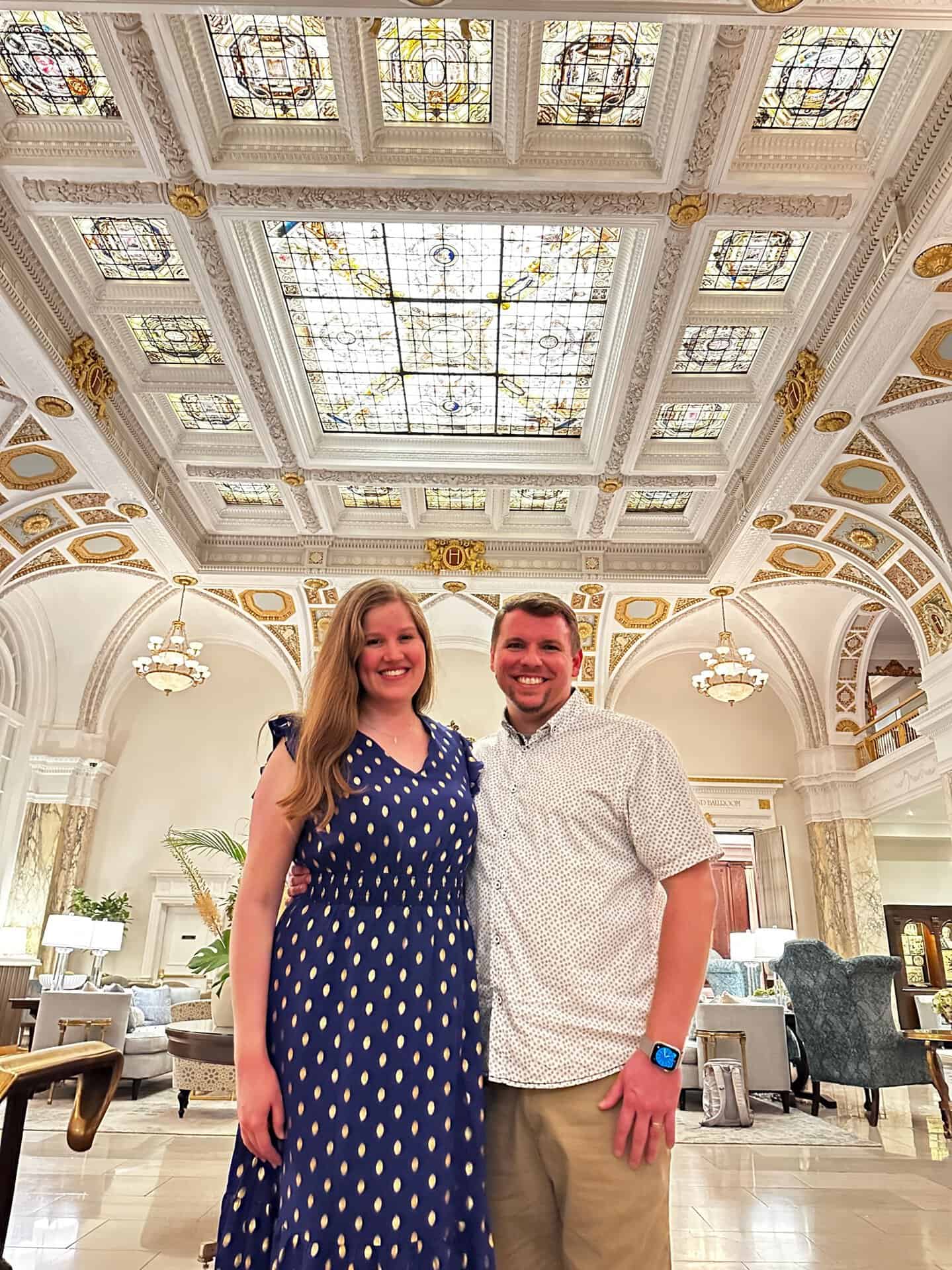 A smiling couple stands in an elegant Nashville room with ornate ceilings and stained glass, featuring intricate designs and chandeliers.
