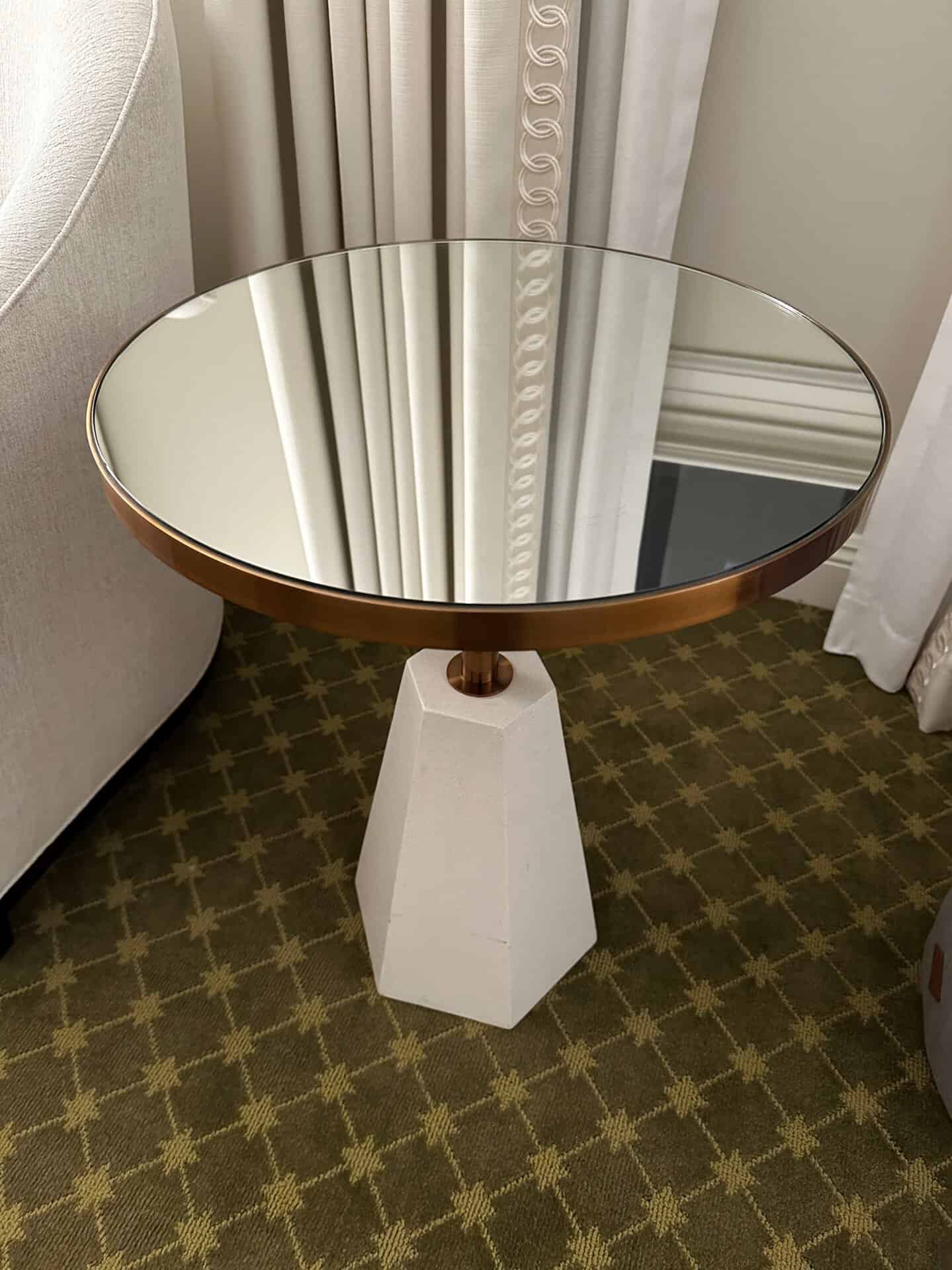A round mirrored side table with a bronze rim stands on a geometric white base, echoing the vibrant style of Nashville. It is placed on a patterned carpet beside curtains.