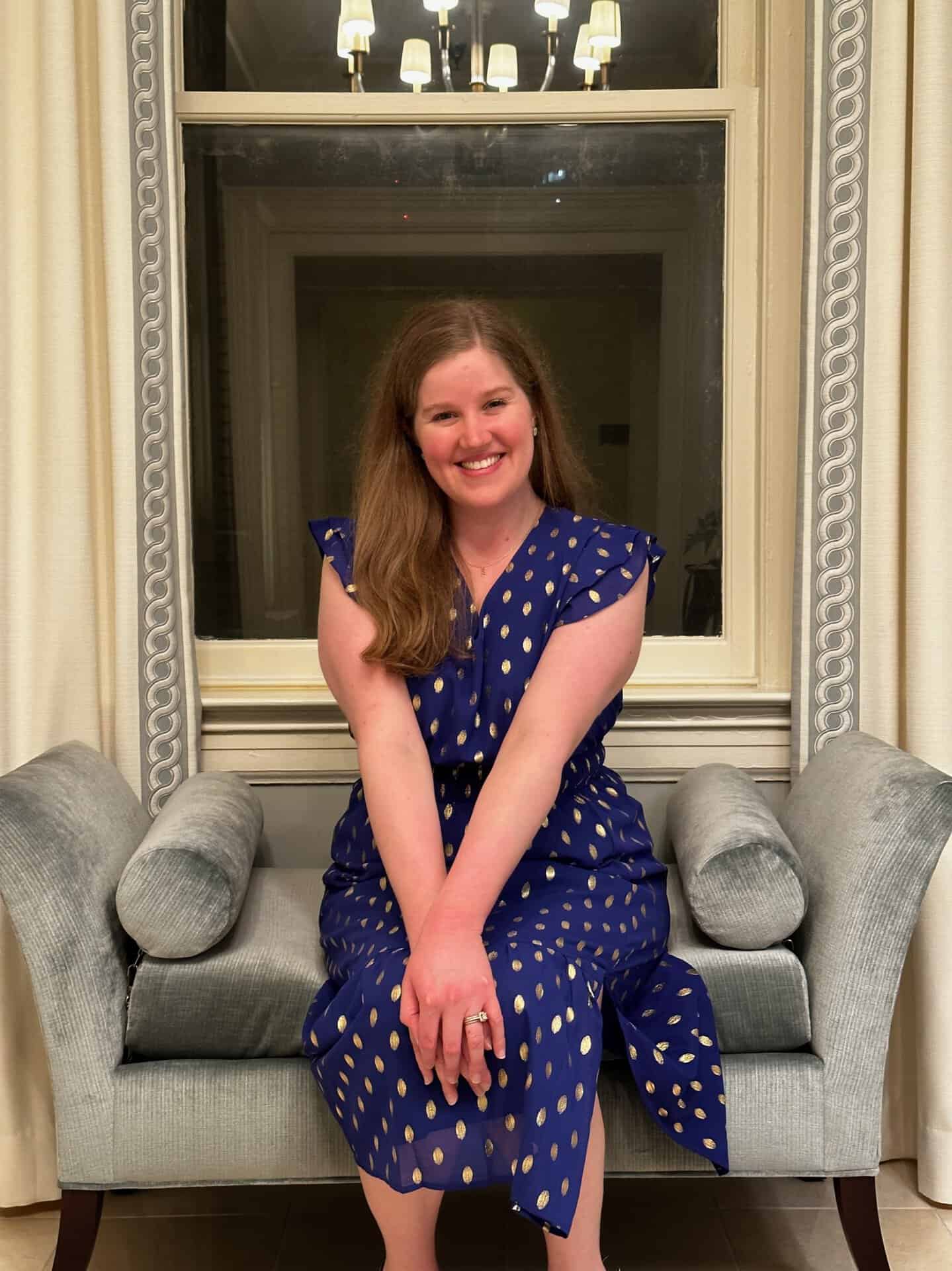 A woman in a blue polka-dotted dress sits on a gray velvet couch, smiling at the camera with Nashville's skyline gleaming through the large window behind her.