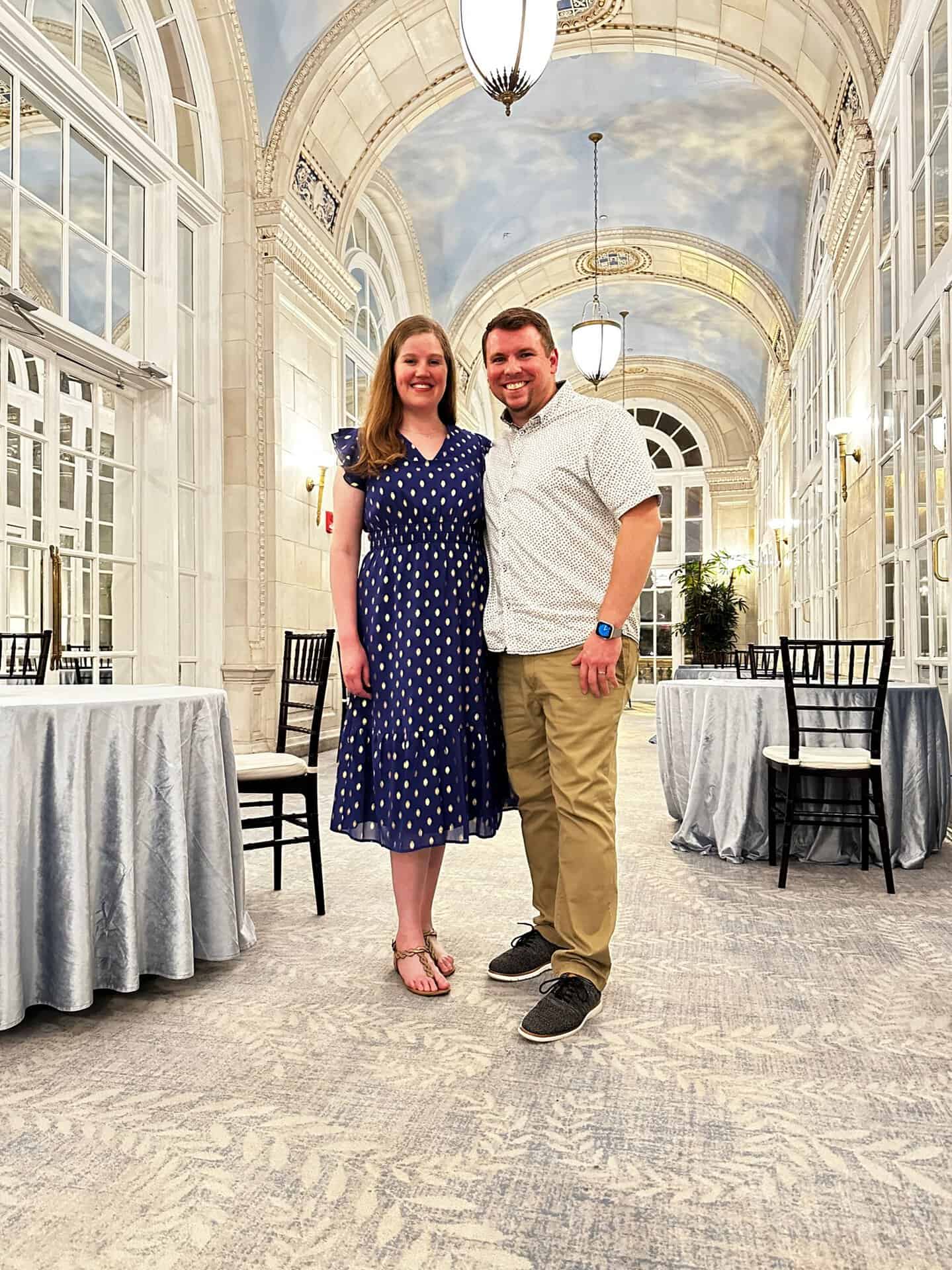 A man and woman stand in an elegant Nashville hallway with arched ceilings and large windows. The woman is in a blue dress with white polka dots, while the man wears a white shirt and khaki pants.
