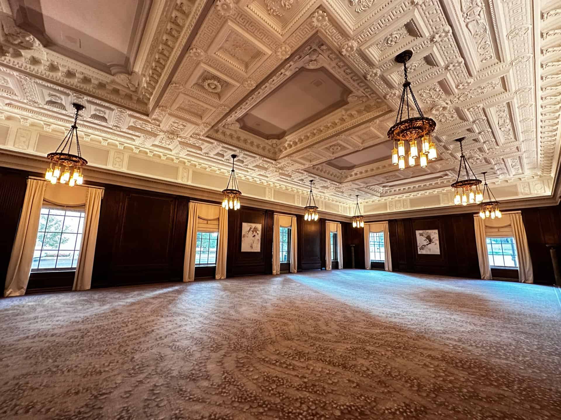 Elegant ballroom in Nashville with an ornate coffered ceiling, chandeliers, large windows draped in curtains, and patterned carpet. The walls showcase two framed artworks, with one door visible.