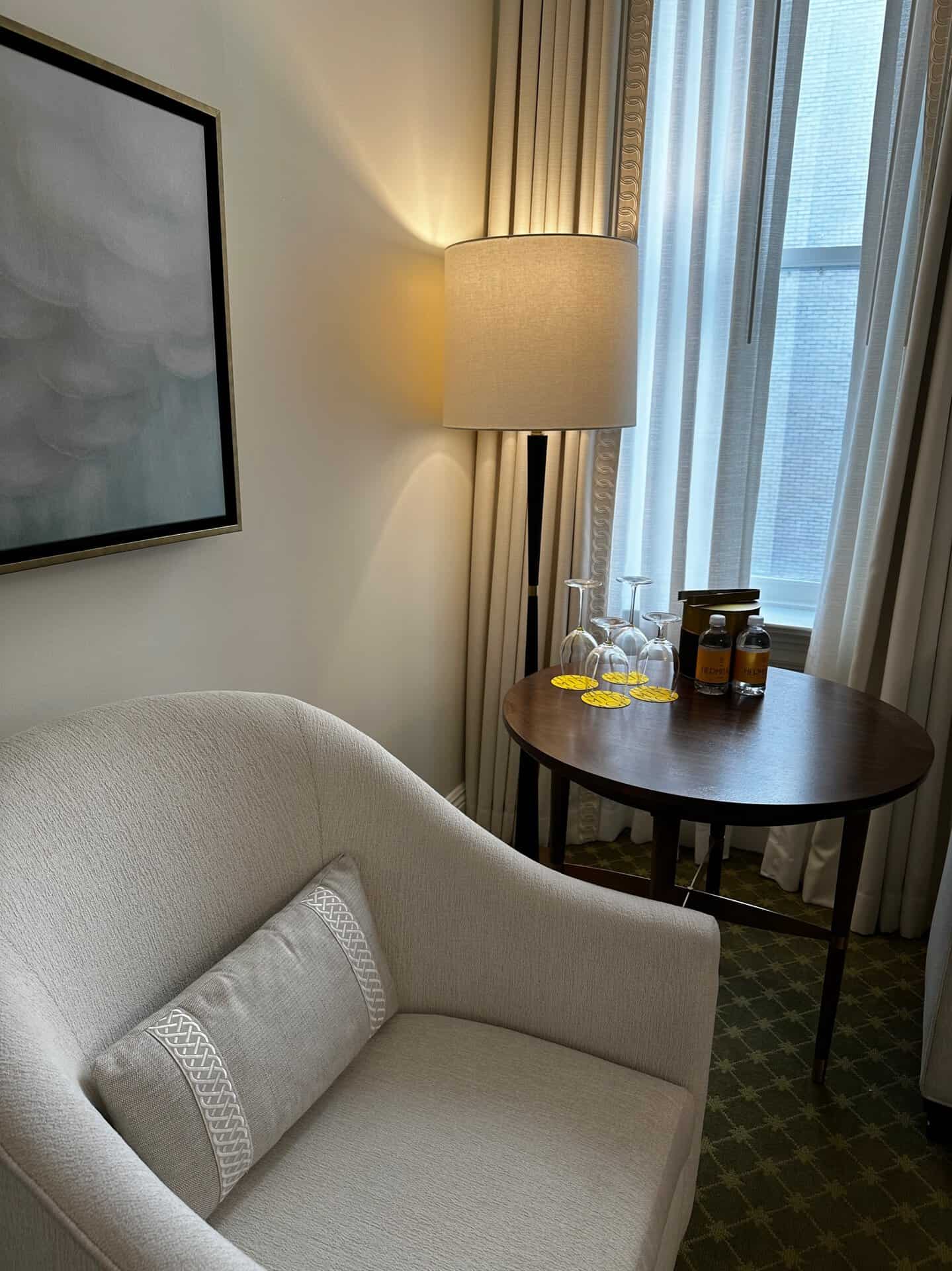 A cozy Nashville nook features a beige armchair, a pillow, and a side table holding three glasses and bottles. A lamp emits warm light beside a window with sheer curtains, creating the perfect Southern comfort vibe.