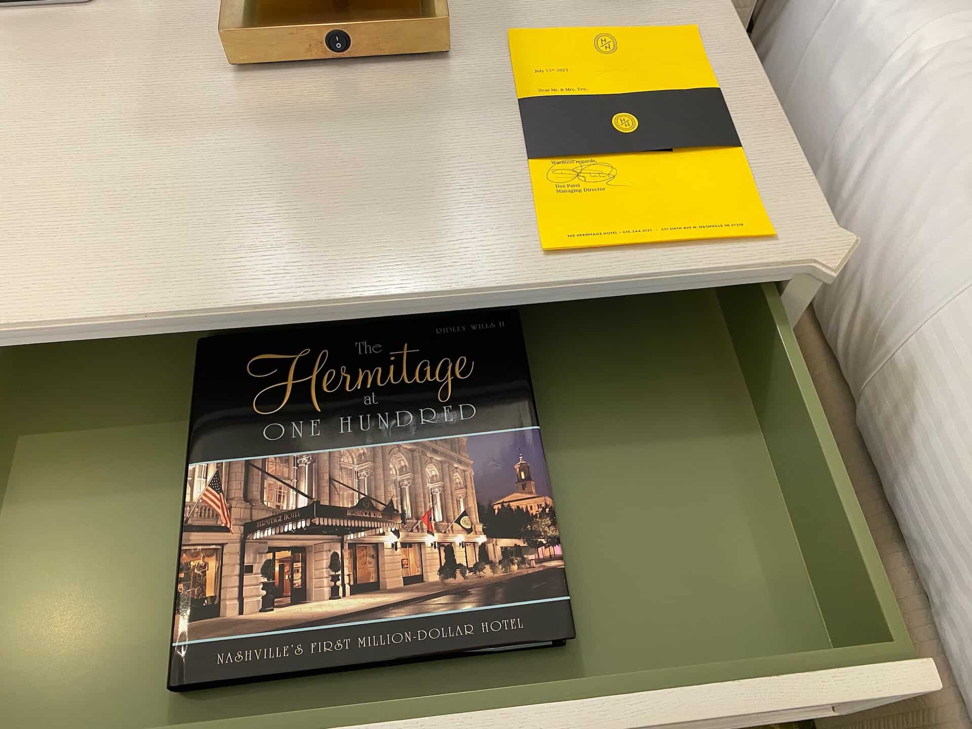 A book titled "The Hermitage at One Hundred" lies in a drawer under a wooden desk, its pages whispering tales from Nashville's storied past. On the desk, a yellow folder with black binding waits patiently for its next reader.
