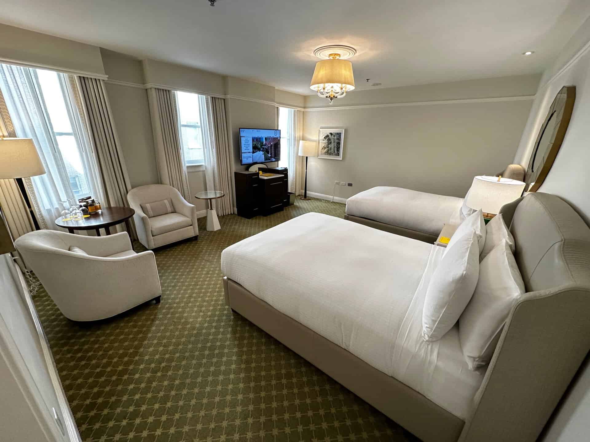 This Nashville hotel room features two beds, a TV, cozy armchairs, and a convenient table. Enjoy the natural light streaming through large windows adorned with curtains. The ambiance is enhanced by a patterned carpet and an elegant ceiling light fixture.