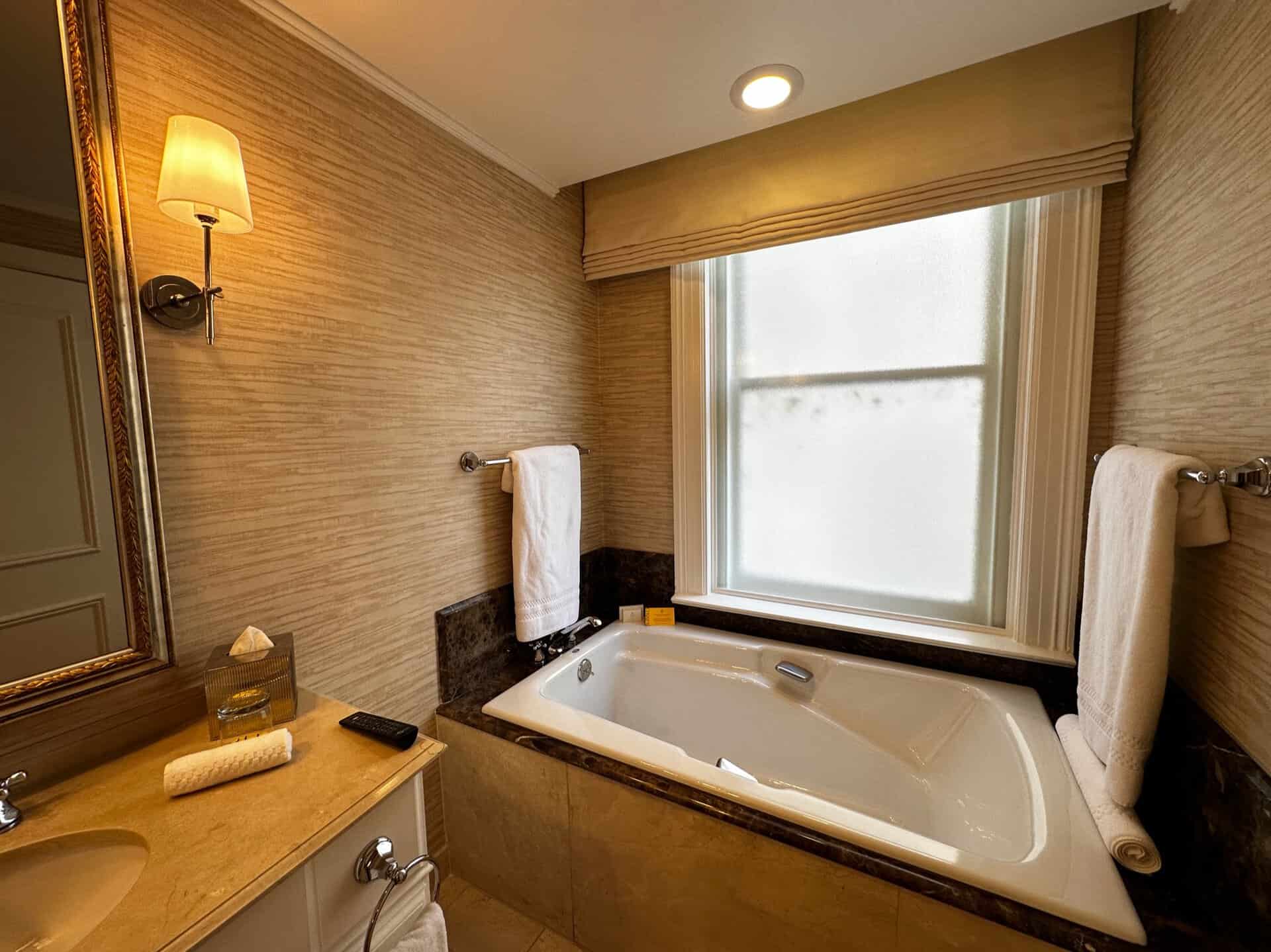 A Nashville-inspired bathroom with a white bathtub under a frosted window, beige walls, and a single wall light. It features a towel rack and a counter adorned with a tissue box and folded towel, embodying the subtle charm of Southern elegance.