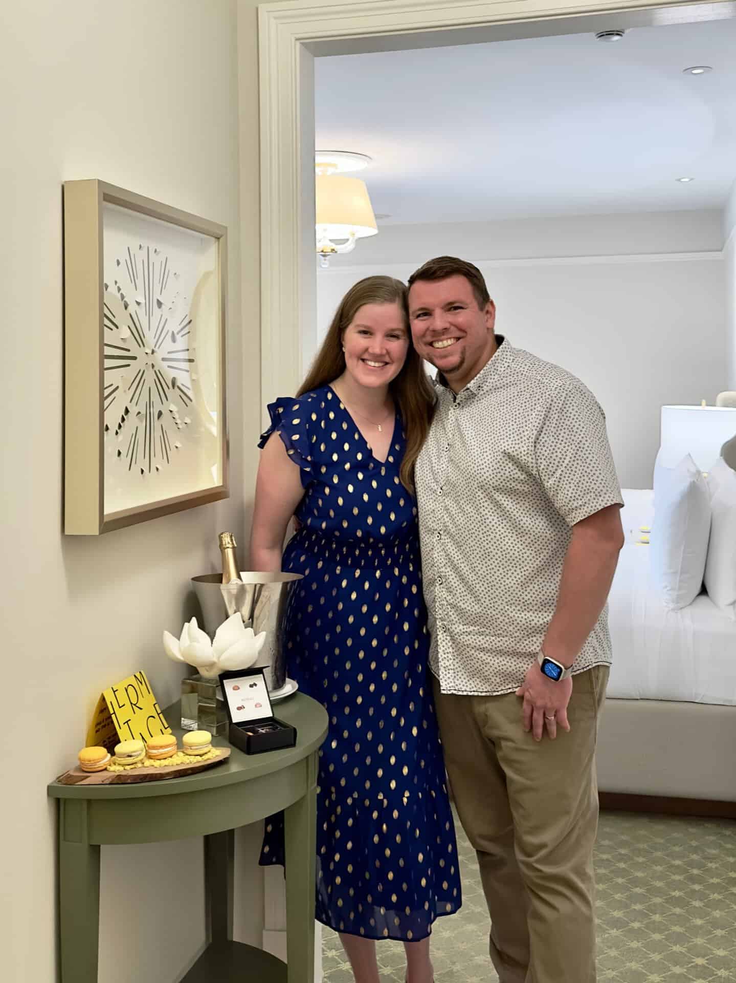 A smiling couple stands next to a small table with champagne and desserts in a cozy Nashville hotel room. A sign with "Merry X-Mas!" is on the table.