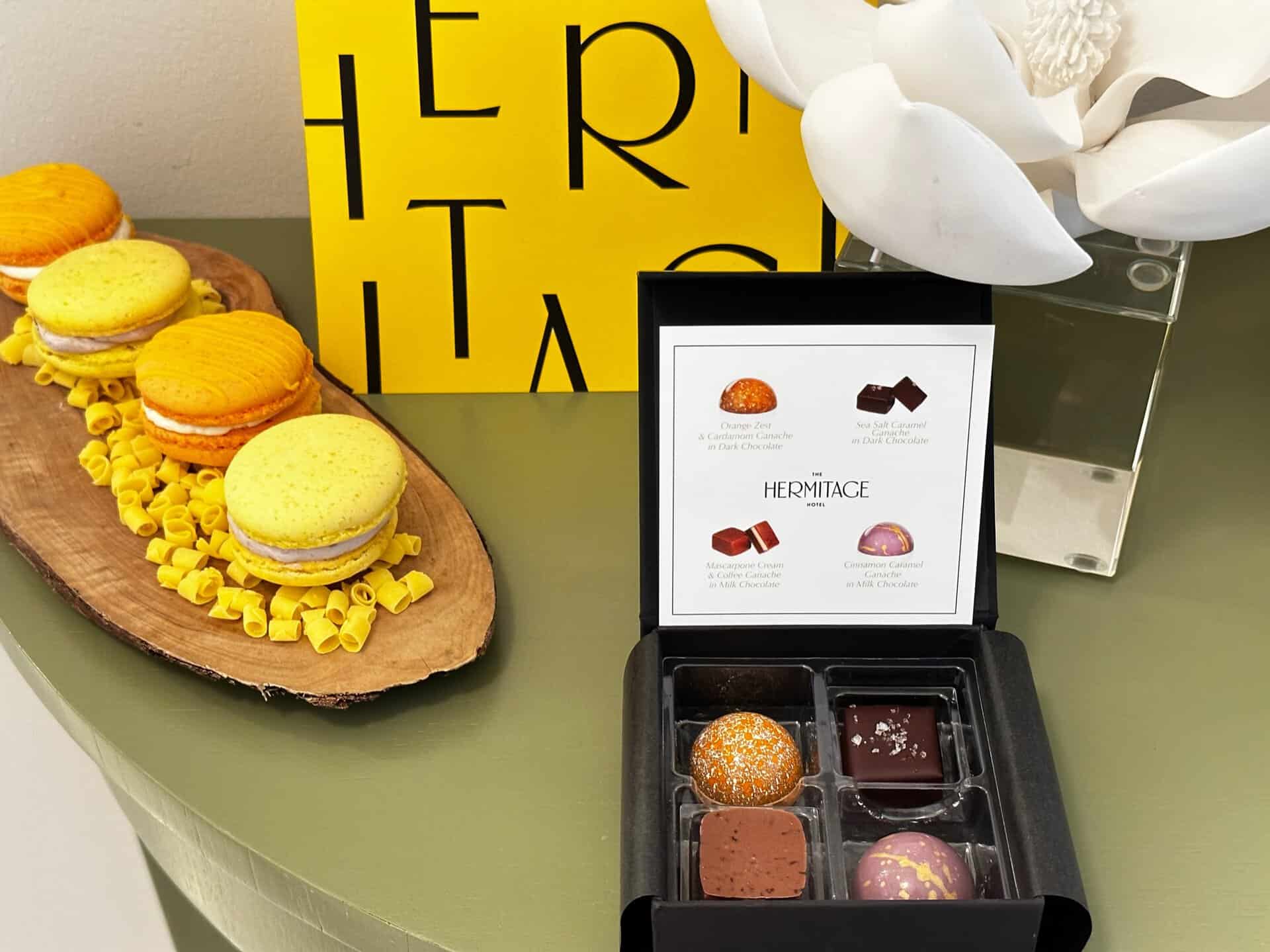 A display with colorful macarons and a box of assorted chocolates sits on a green table, evoking the charm of Nashville. A white flower and a yellow "HERMITAGE" sign subtly accentuate the background.
