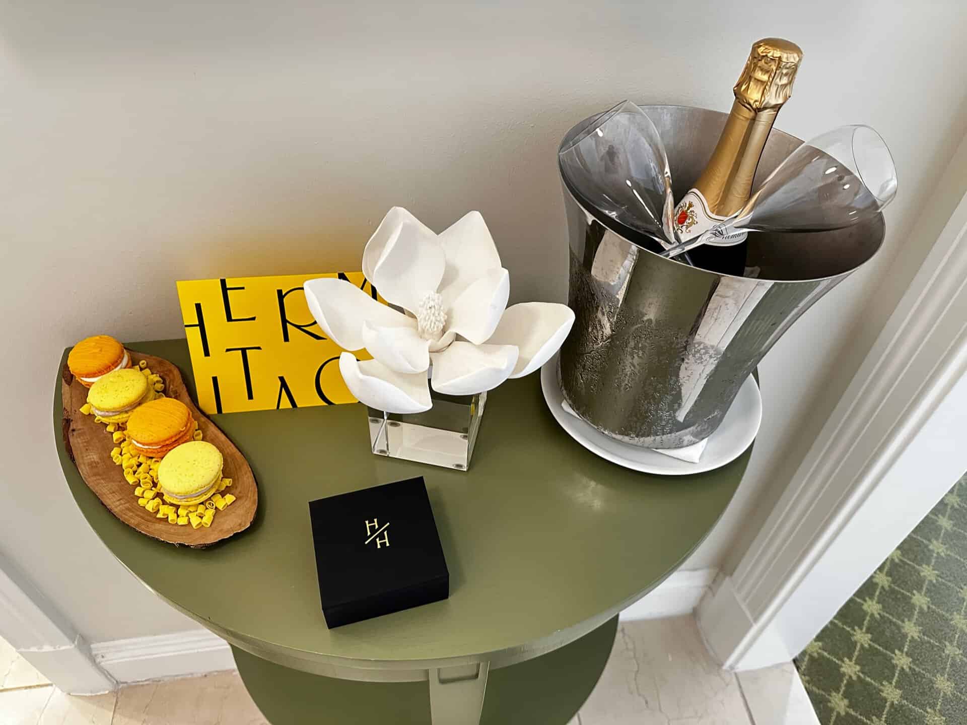 Champagne in an ice bucket, a magnolia flower in a vase, a small black box, and macarons on a wooden plate are arranged on a green table, evoking the charm of Nashville.