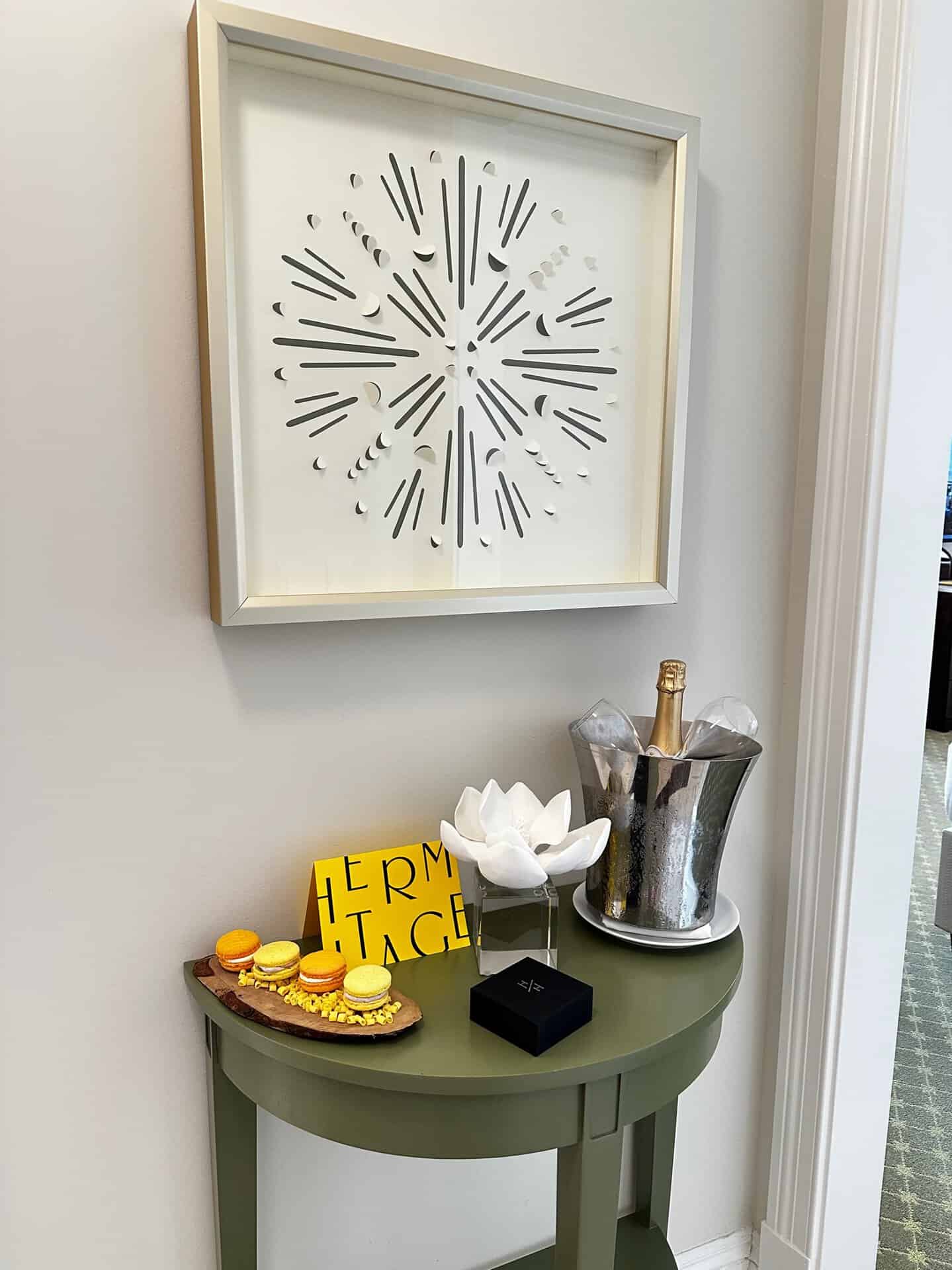 A decorative wall art piece hangs above a green table reminiscent of Nashville's vibrant charm, complete with a champagne bucket, a small box, a white flower sculpture, a yellow sign, and a wooden tray adorned with macarons.