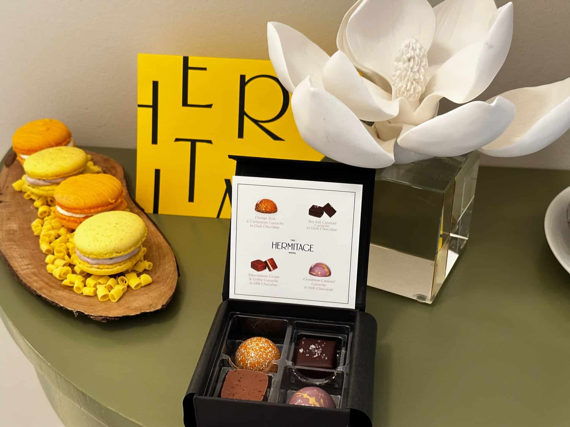 An open box of four assorted chocolates is displayed on a green surface, alongside yellow macarons and a decorative white flower in a glass vase. A yellow card with "HERMITAGE" text nods to Nashville's charm.