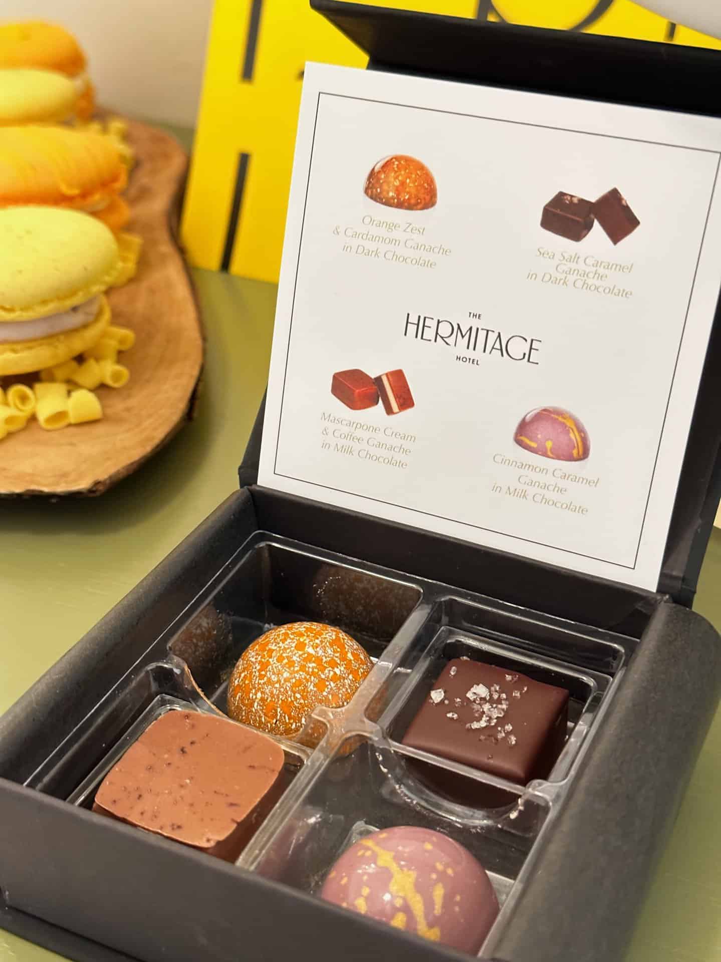 A box of assorted chocolates comes with a menu card featuring flavors like orange zest, sea salt caramel, macaroon cream, and cinnamon caramel. In the background, a plate with macarons adds an inviting touch reminiscent of Nashville's vibrant culinary scene.