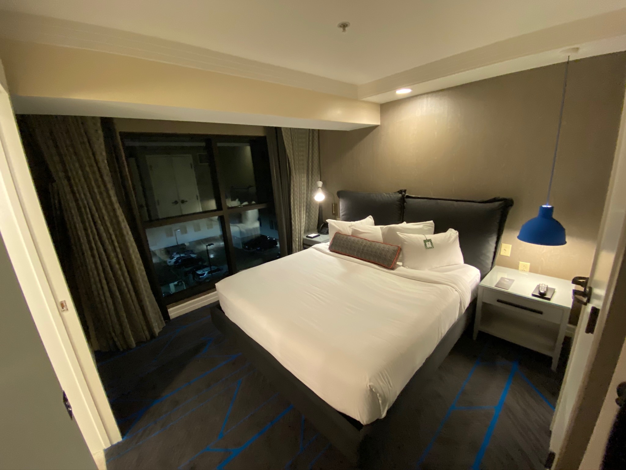A modern hotel room with a neatly made bed, two nightstands, a blue hanging lamp, and a large window with dark curtains.