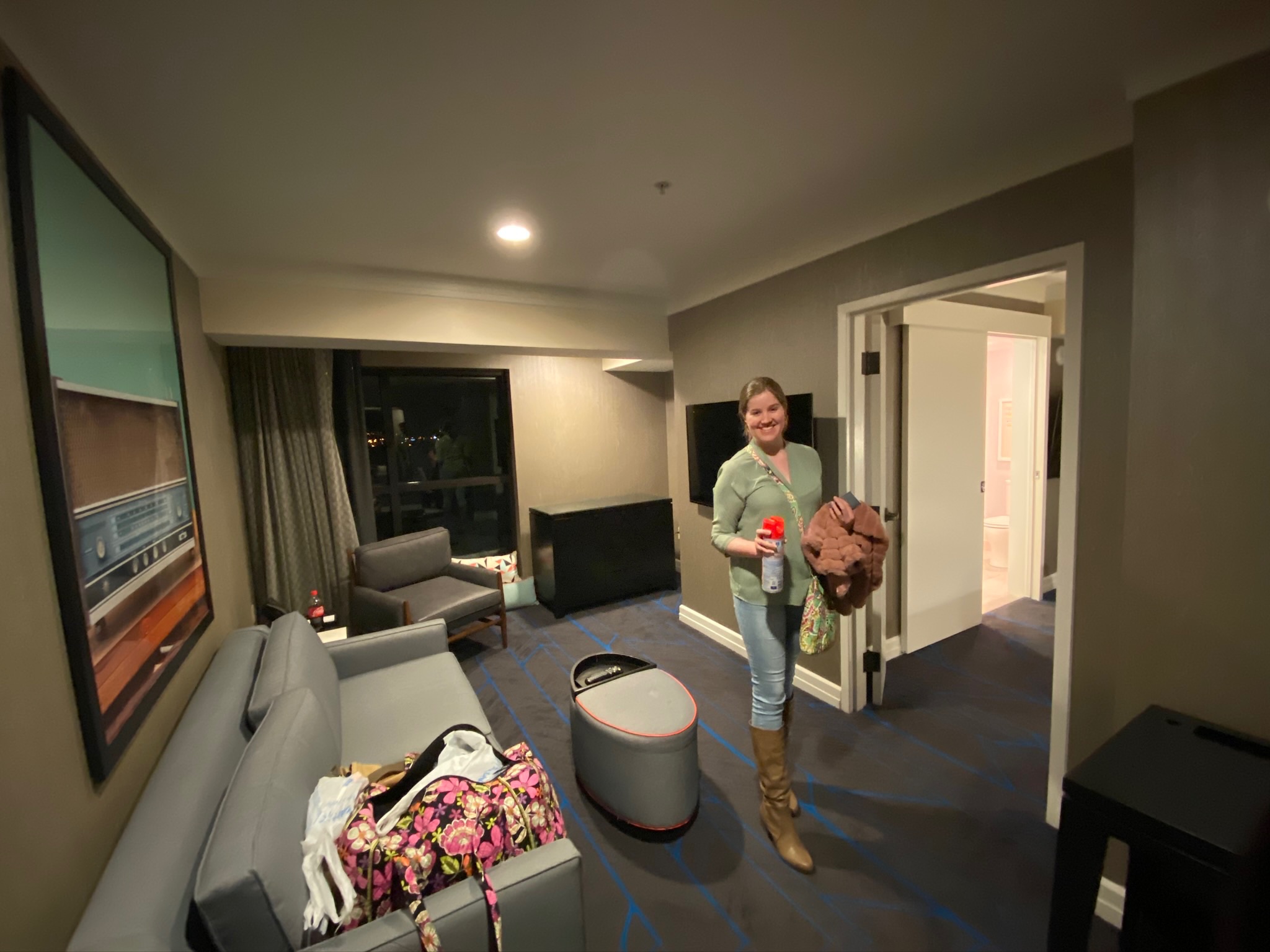 A woman stands smiling in a hotel room holding a drink and a jacket. The room has a sofa, bags on the floor, and an open door leading to another room.