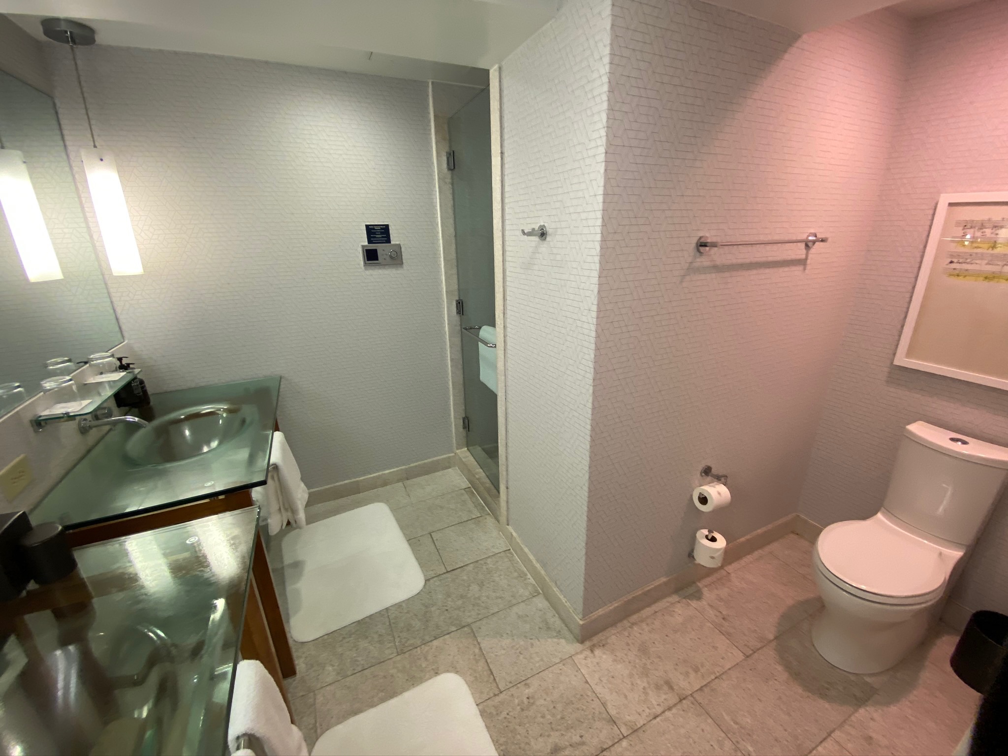 Modern bathroom with a glass sink, wall mirror, towels, walk-in shower, toilet, toilet paper rolls, towel rack, and framed artwork on a tiled wall.
