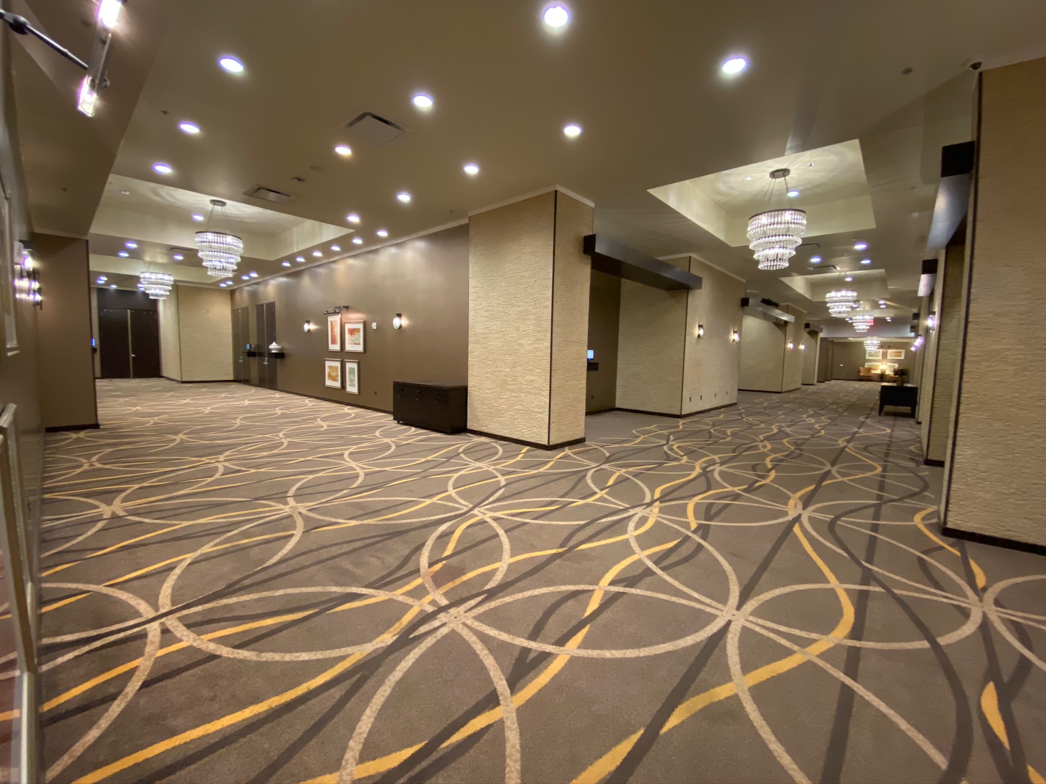 Wide, empty hotel hallway with patterned carpet, overhead lighting, chandeliers, and framed art on the walls.