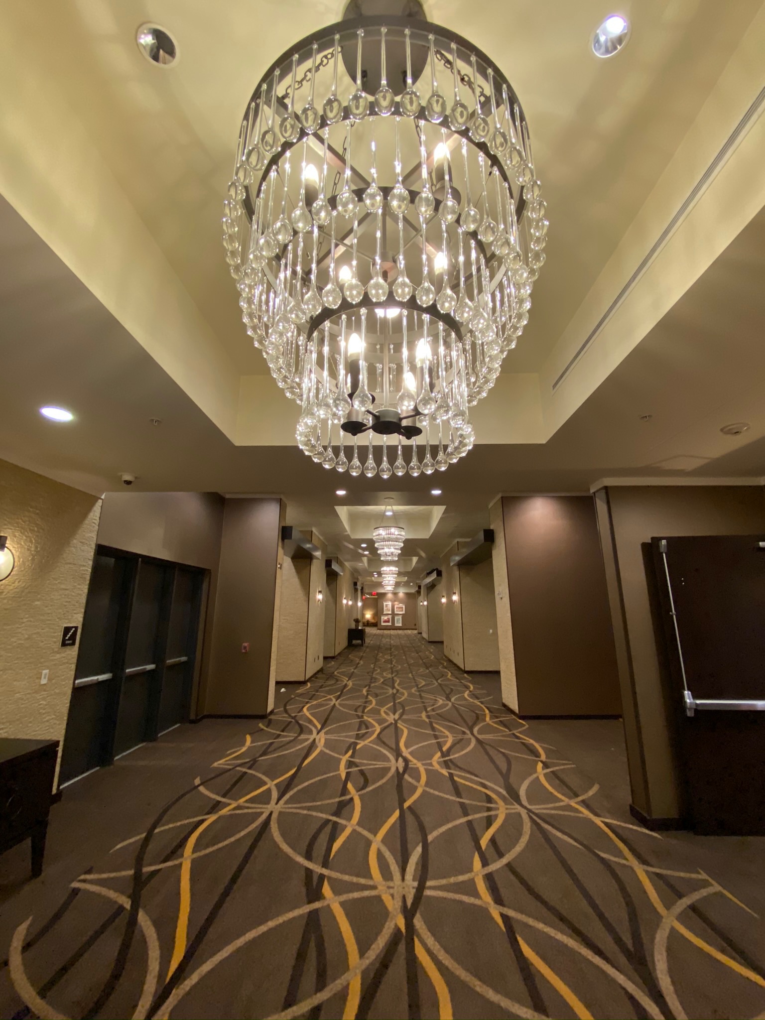 A hotel hallway with patterned carpet, dark wall panels, and three large crystal chandeliers hanging from the ceiling.