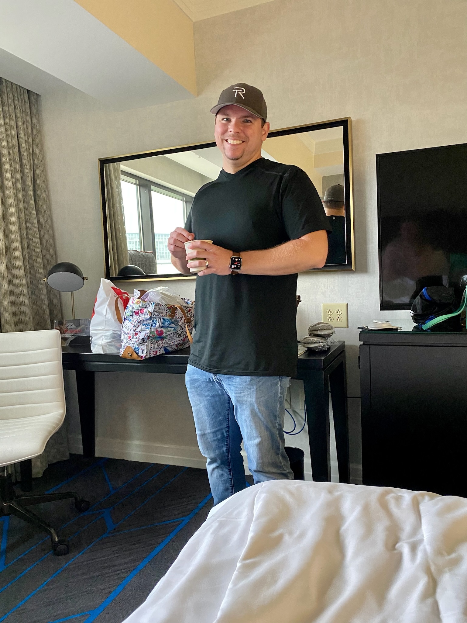 A man in a black shirt, jeans, and a gray cap stands in a hotel room, holding a cup. A television, desk, and chair are visible in the background.