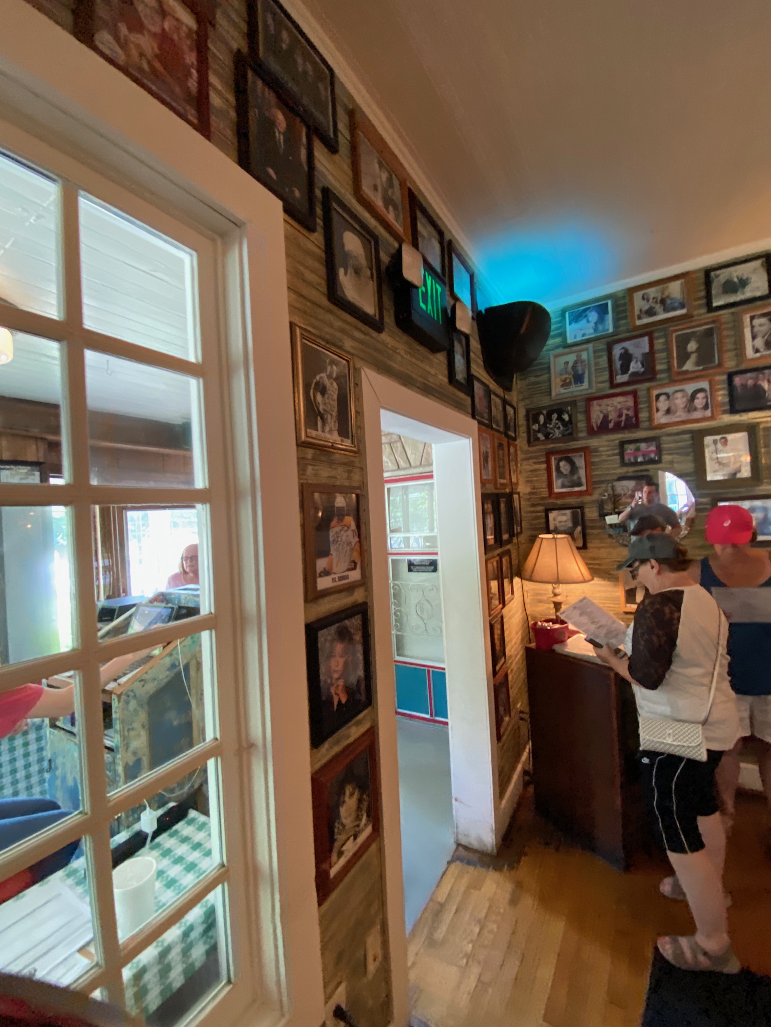 People stand near a reception area in a restaurant with wood floors and walls covered in framed photographs; a lamp sits on a small table by the wall.