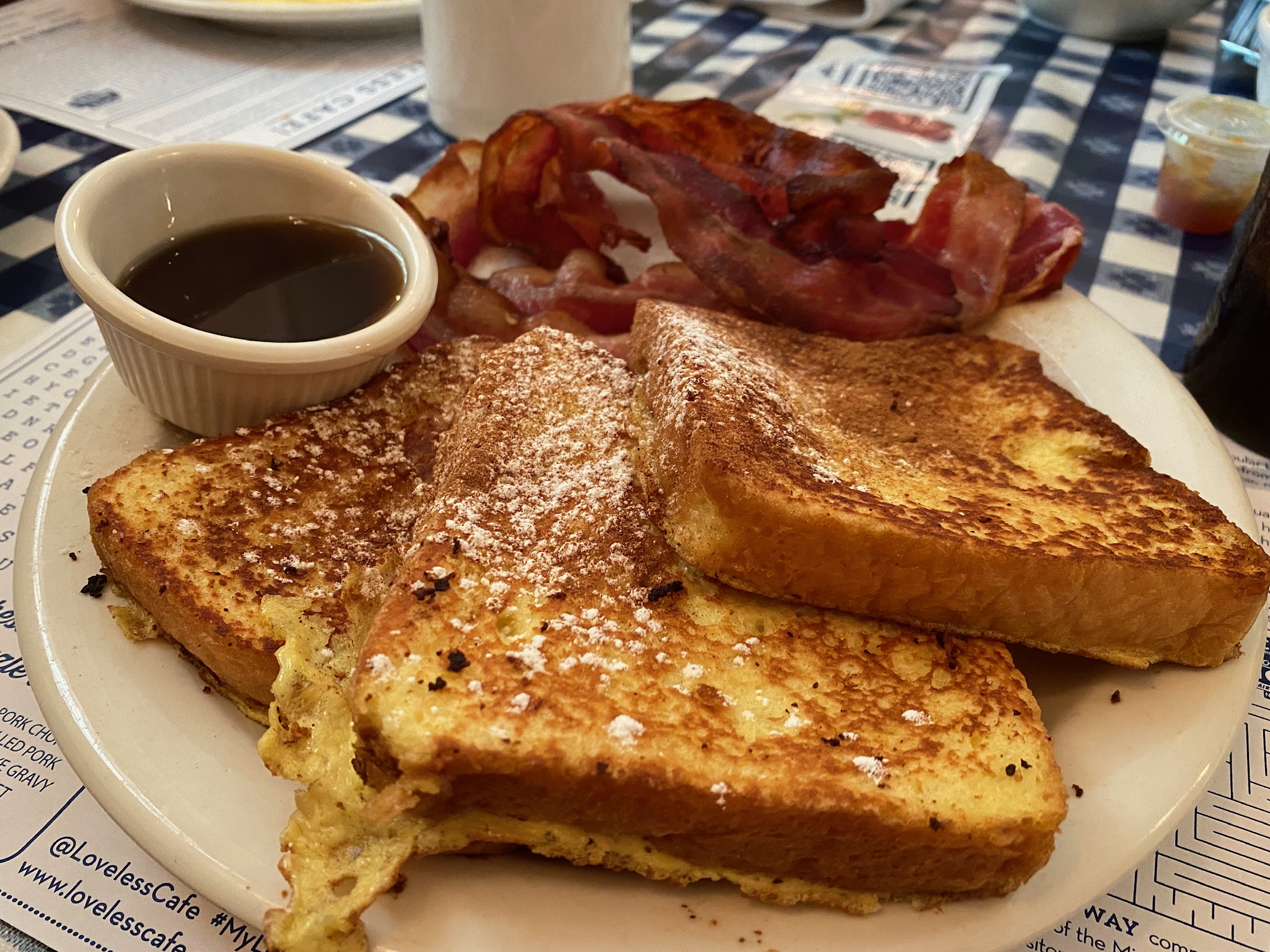 A plate with two slices of French toast, several strips of bacon, and a small cup of syrup on a checkered tablecloth.