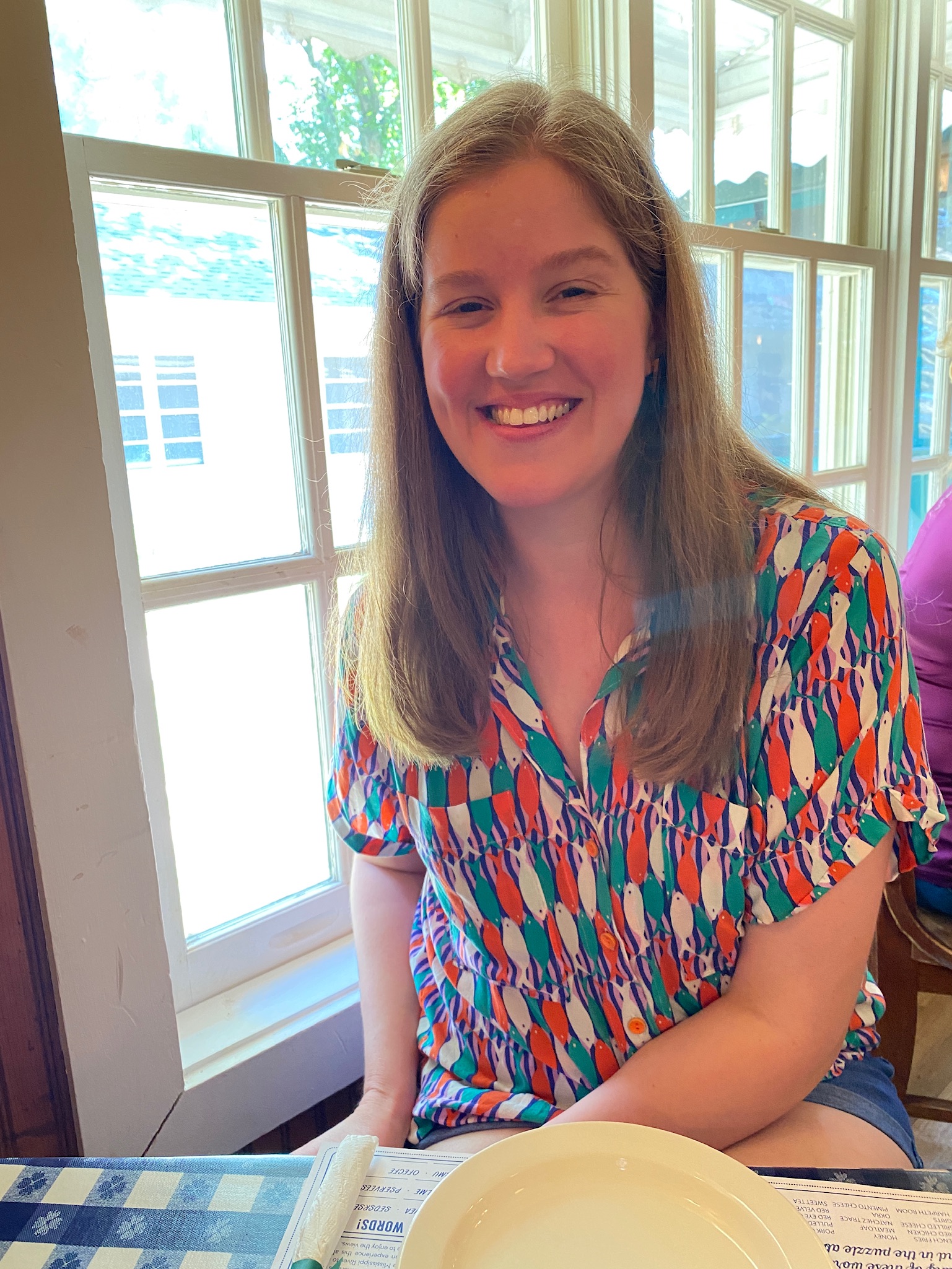 A woman with long brown hair sits at a table by a window, smiling. She wears a colorful patterned shirt and has an empty plate in front of her.