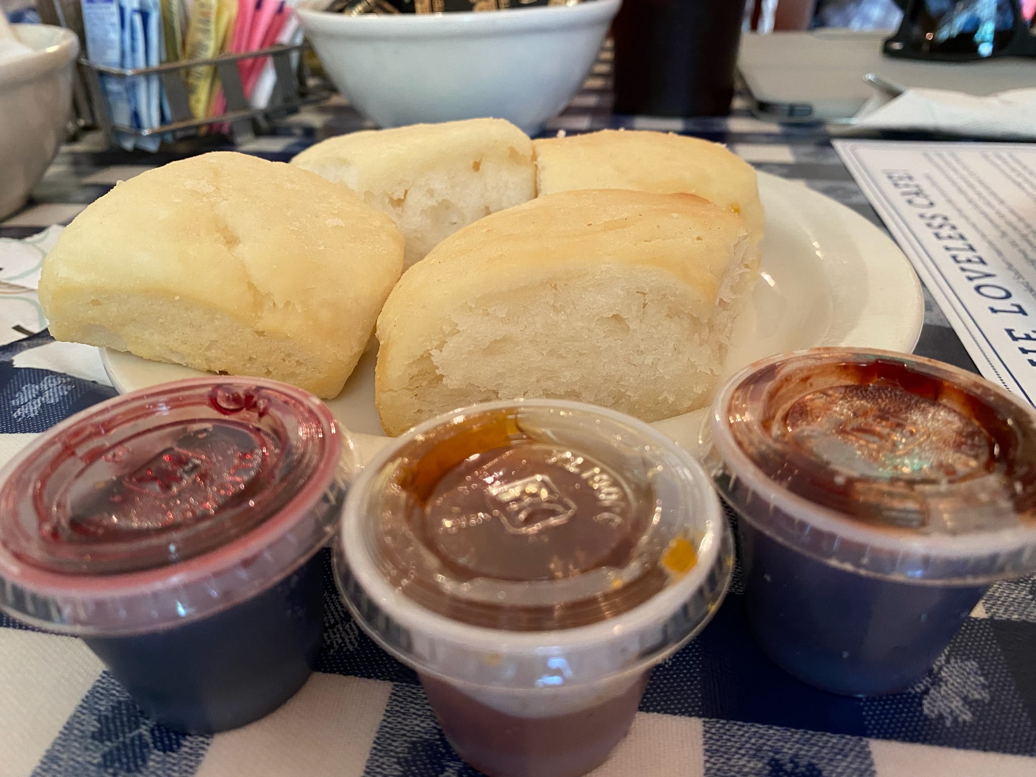 Four bread rolls on a white plate with three small containers of jam or preserves on a blue and white checkered tablecloth.