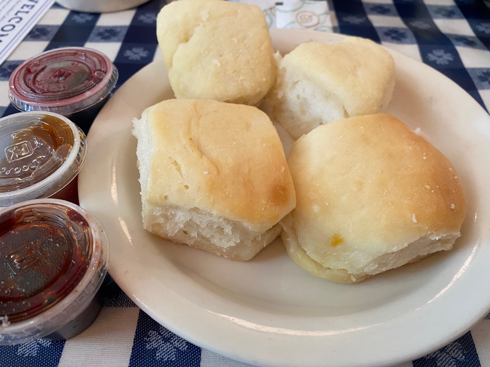 Four plain dinner rolls on a white plate next to three small containers of jam, placed on a blue and white checkered tablecloth.