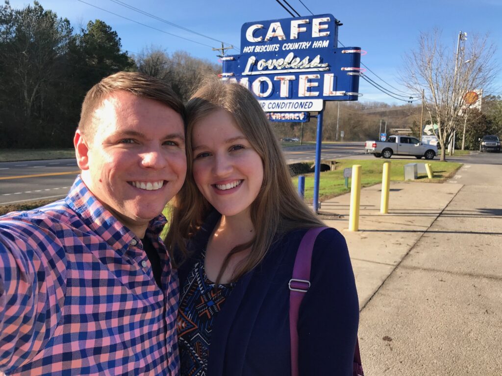 A man and woman smile for a selfie in front of the blue Loveless Cafe and Motel sign on a sunny day.