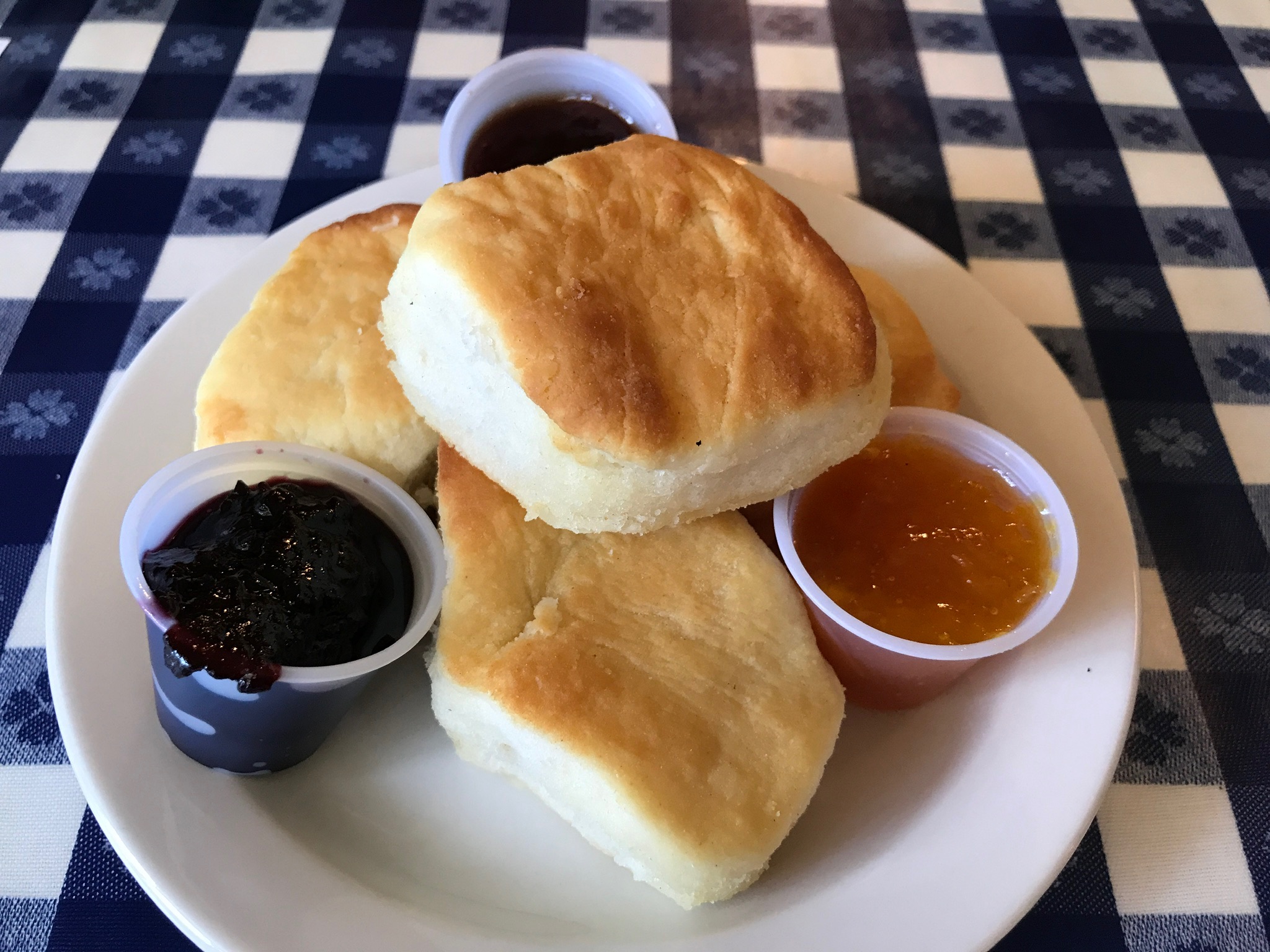 Three biscuits on a white plate with three small cups of various fruit jams, set on a blue and white checkered tablecloth.