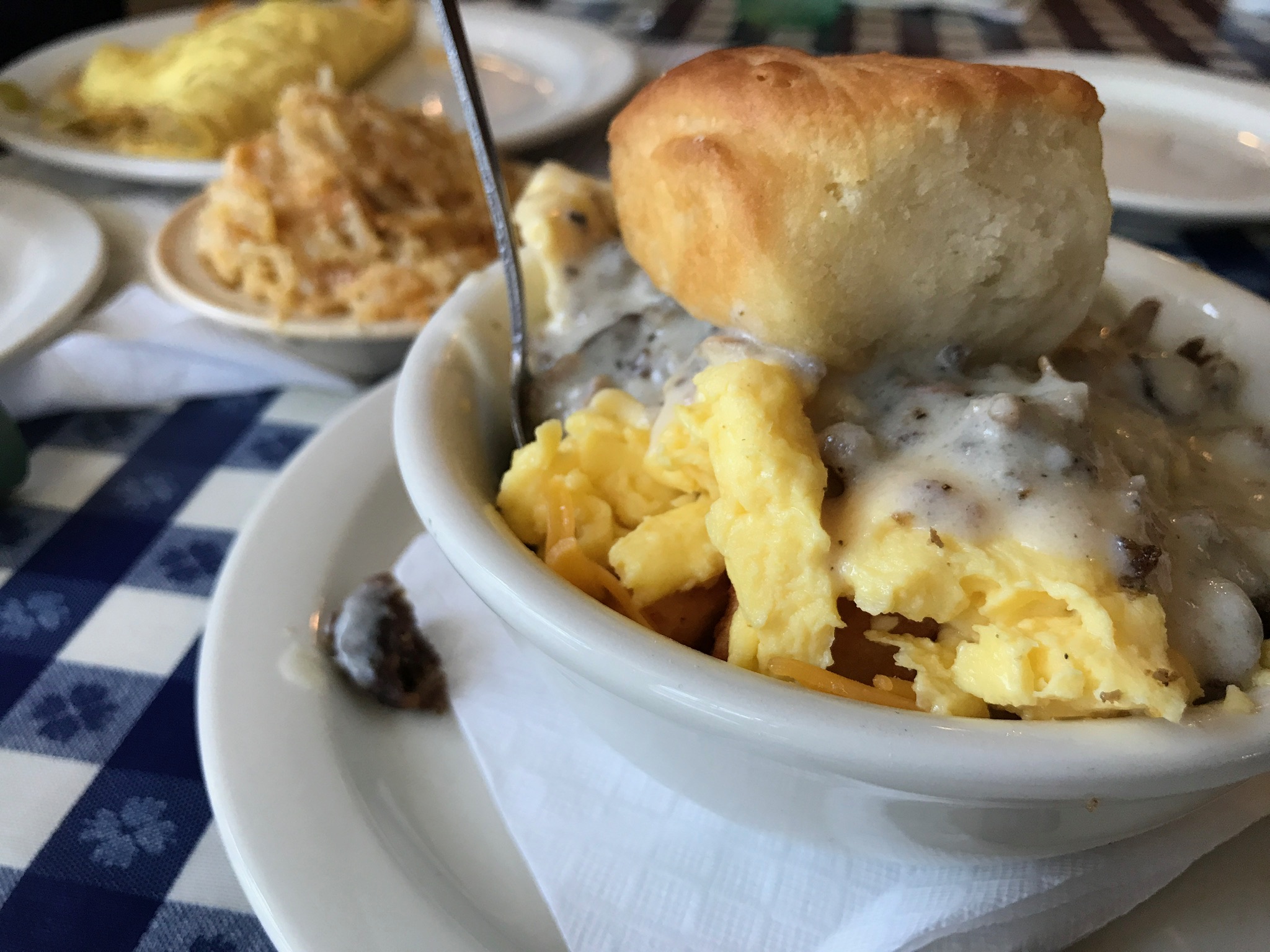 A bowl of scrambled eggs topped with sausage gravy and a biscuit, with a plate of macaroni and cheese and an omelet in the background on a checkered tablecloth.