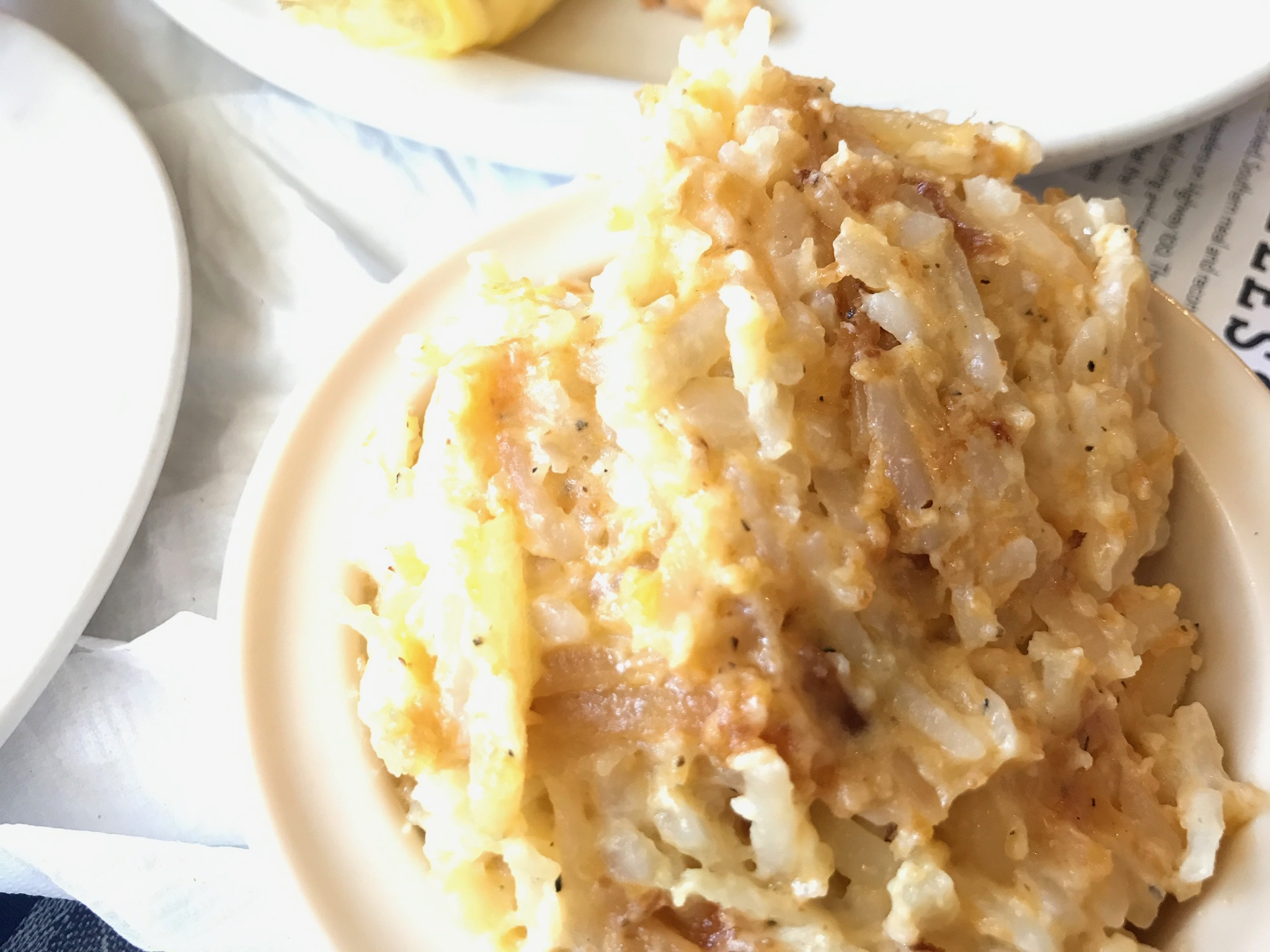 A close-up of a serving of cheesy hash brown casserole on a white plate, with parts of other plates visible in the background.