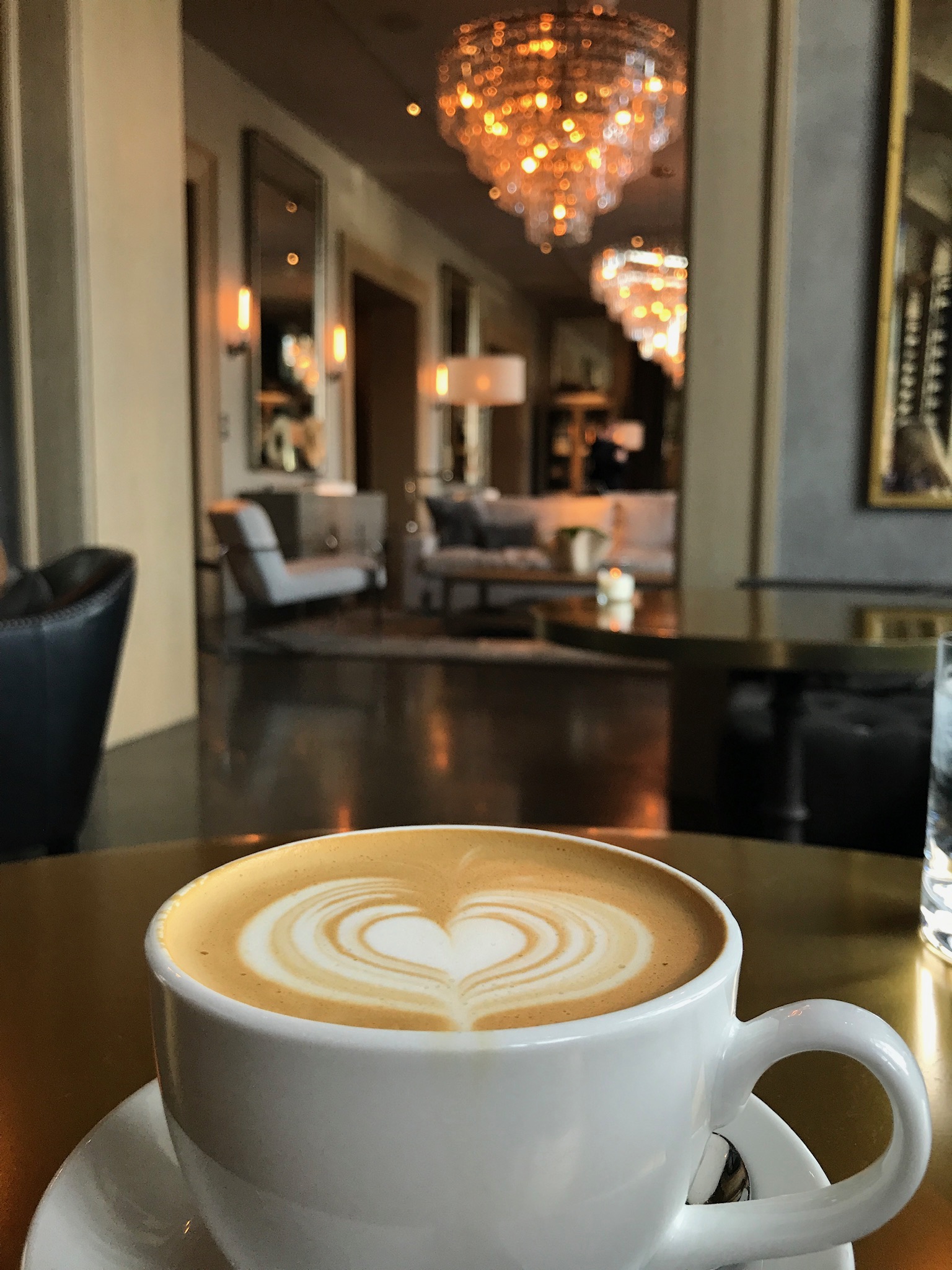A cup of latte with heart-shaped foam art sits on a table in a modern, well-lit lounge with chandeliers and contemporary furniture in the background.